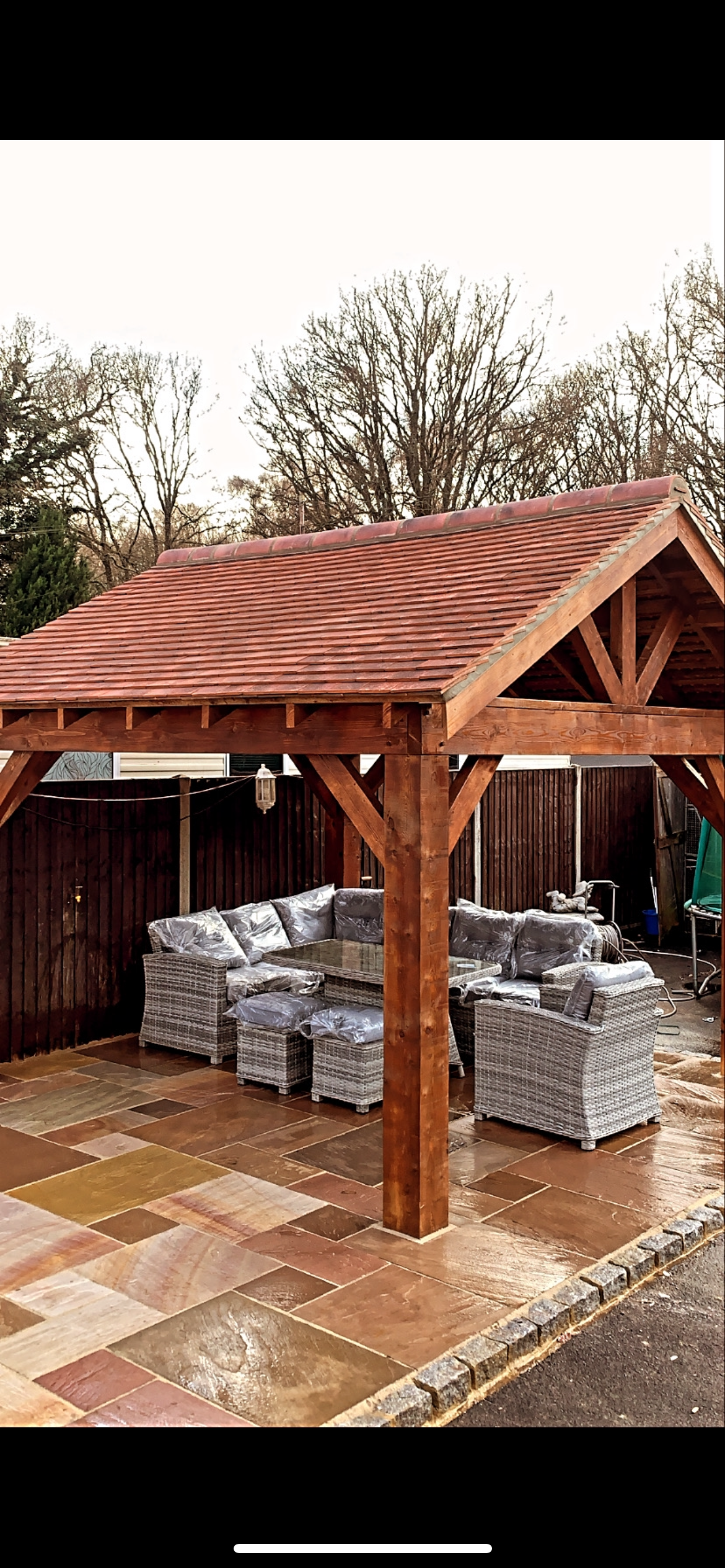 A backyard patio with a wooden pergola covering outdoor wicker furniture, including a table and chairs, on a tiled stone floor, with leafless trees and a fence in the background.