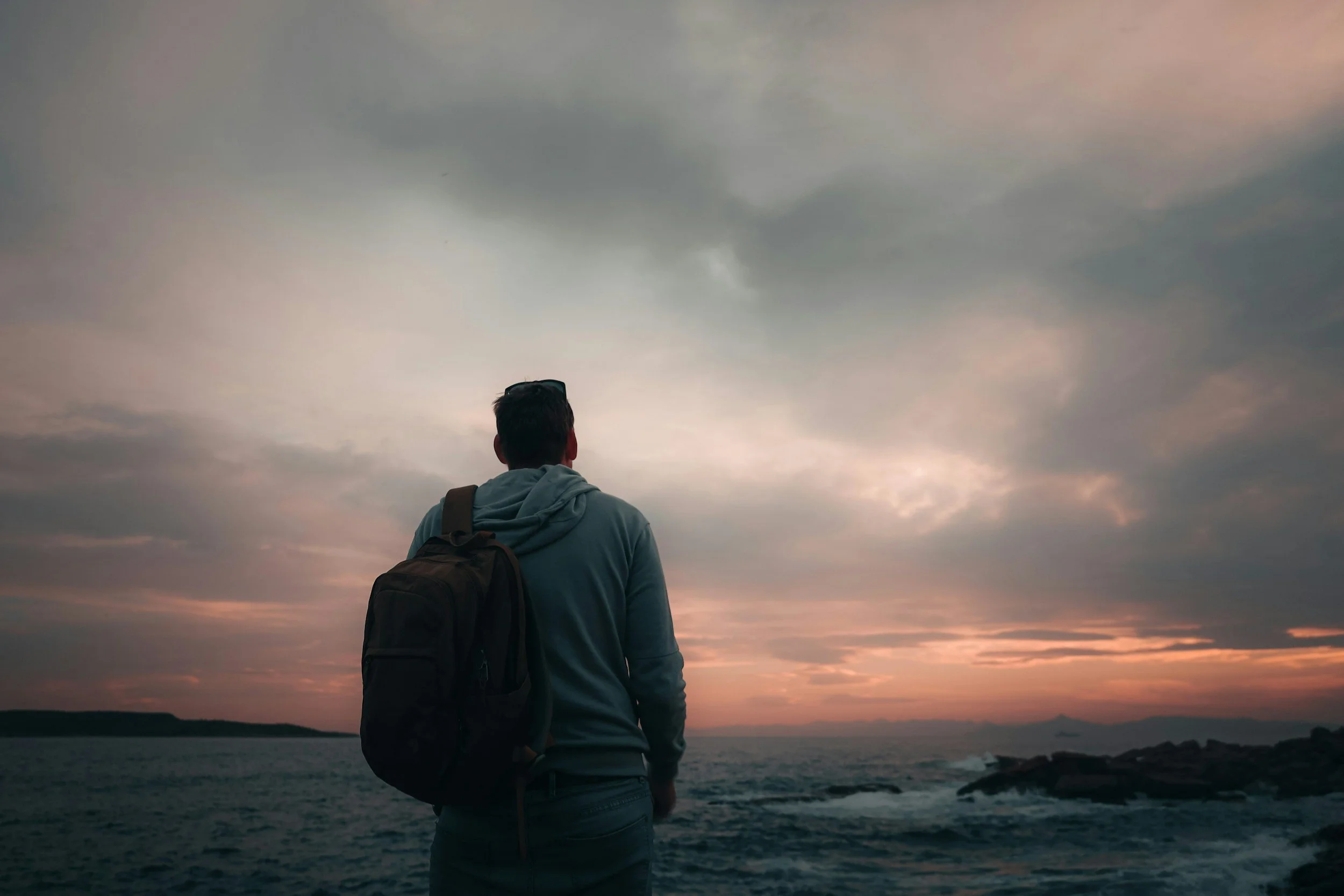 A person with a backpack looking out over a body of water during sunset or dusk, with a cloudy sky overhead.