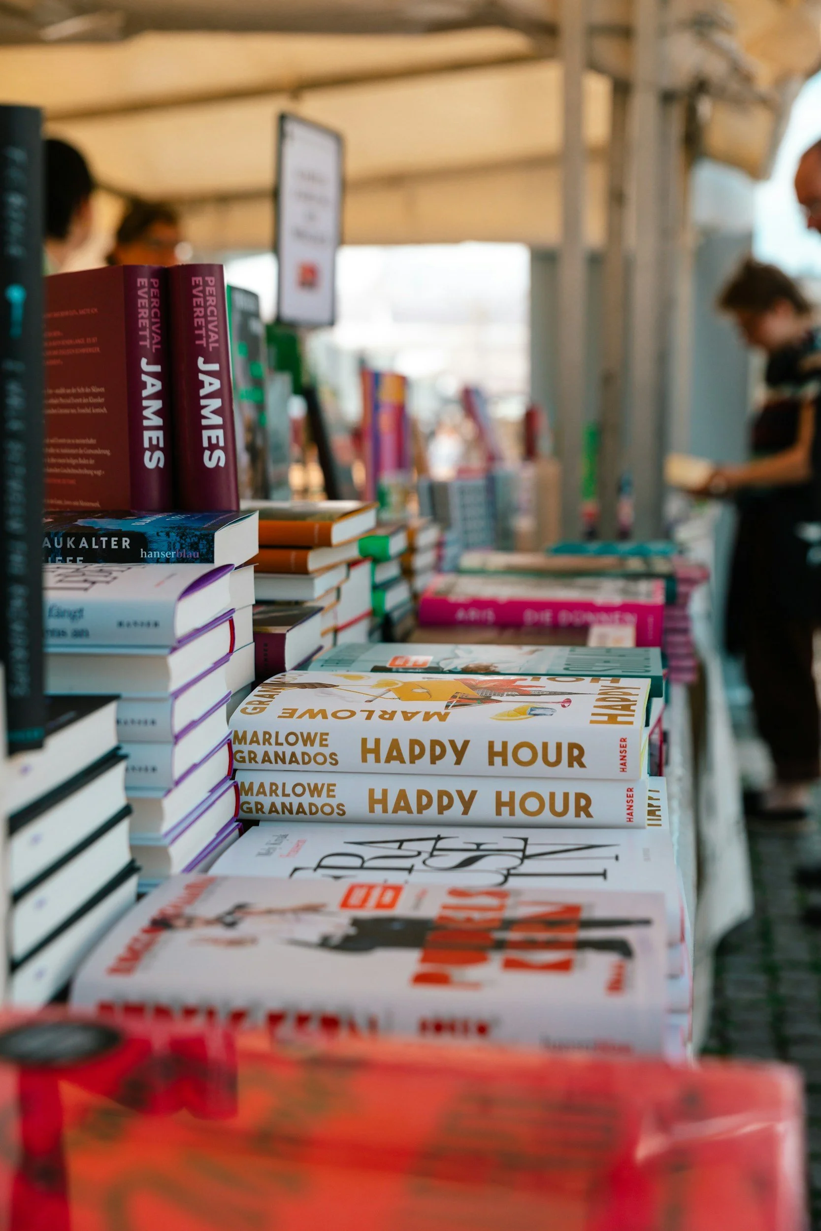 Stack of books on a table with copies of 'Happy Hour' written by Marlowe Granados in the foreground, shelves of books in the background with people browsing.