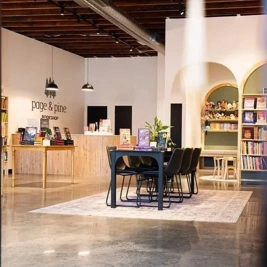 Interior of a bookstore with a central table, chairs, a wooden counter, bookshelves, and a rug.