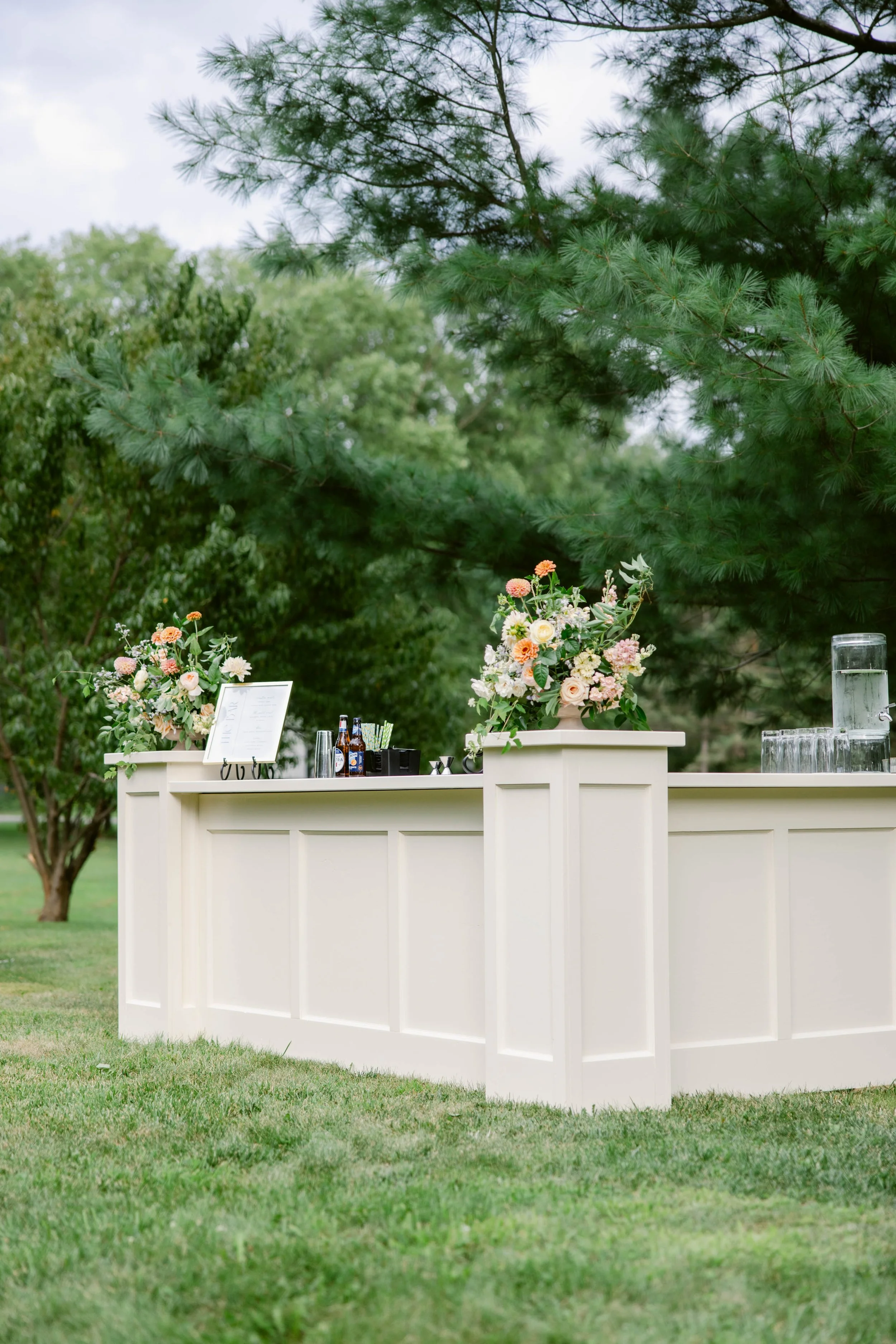 Outdoor bar setup with floral arrangements on a white counter, surrounded by green trees and grass.