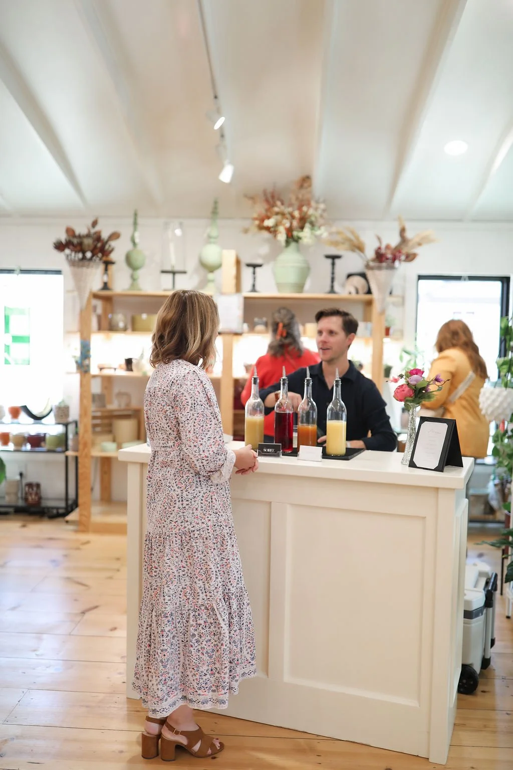 A woman at a checkout counter in a flower shop, talking to a man behind the counter. The shop is decorated with flowers, vases, and other decorative items.