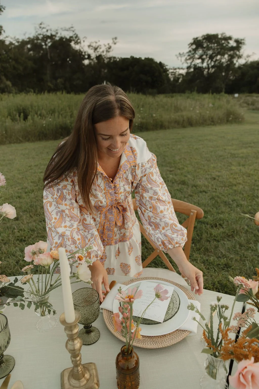 A woman with long brown hair smiling and arranging table settings outdoors with a nature background, surrounded by flowers and candles.
