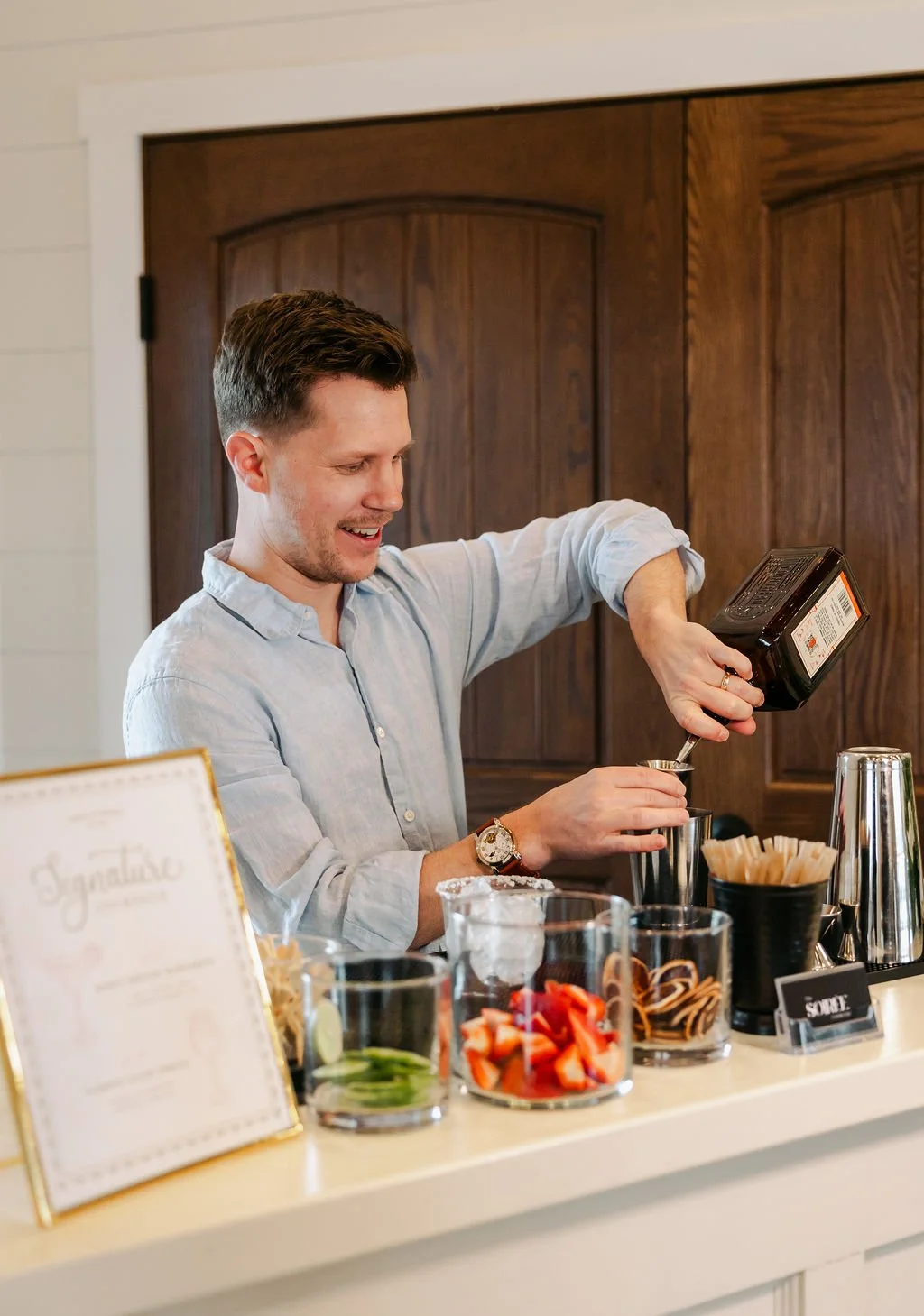 A man in a light blue button-up shirt pouring syrup into a small cup at a bar or cafe counter with various ingredients and containers around him.
