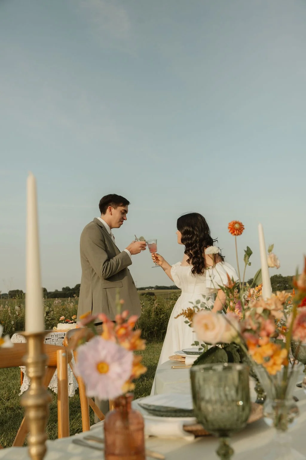 A man and woman in wedding attire standing outdoors at a decorated table, clinking glasses during a celebration or wedding reception.