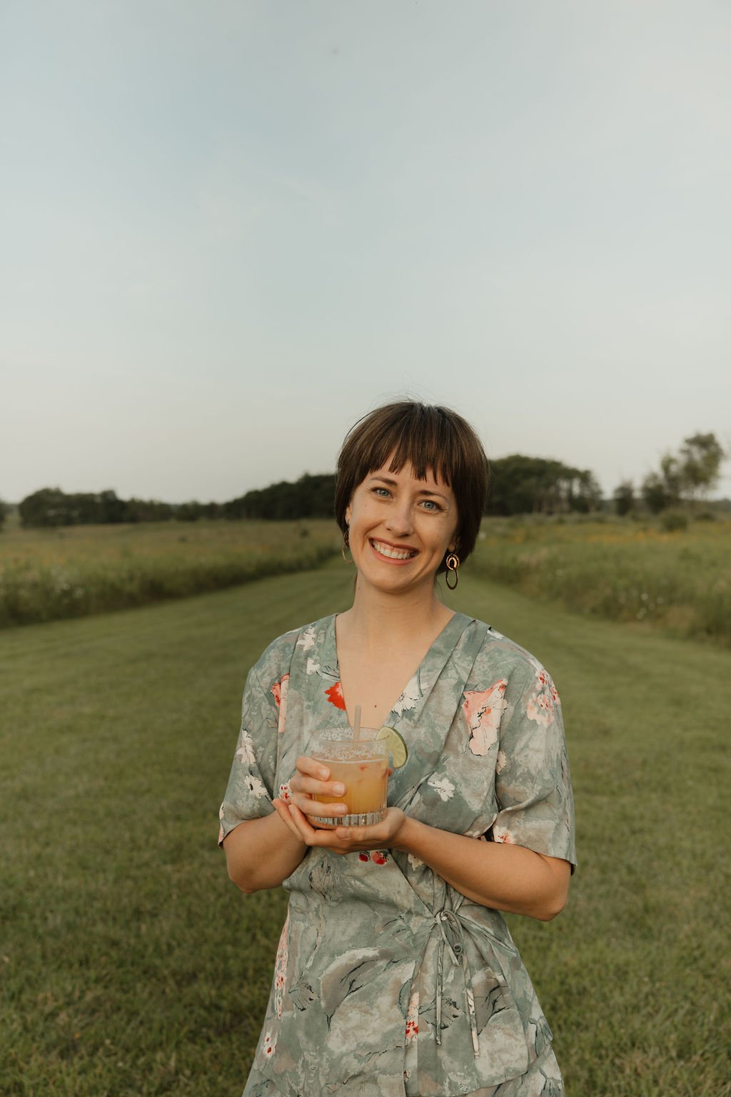 A woman with short brown hair, wearing a floral dress, smiles and holds a drink with a lime wedge in an open field during daytime.