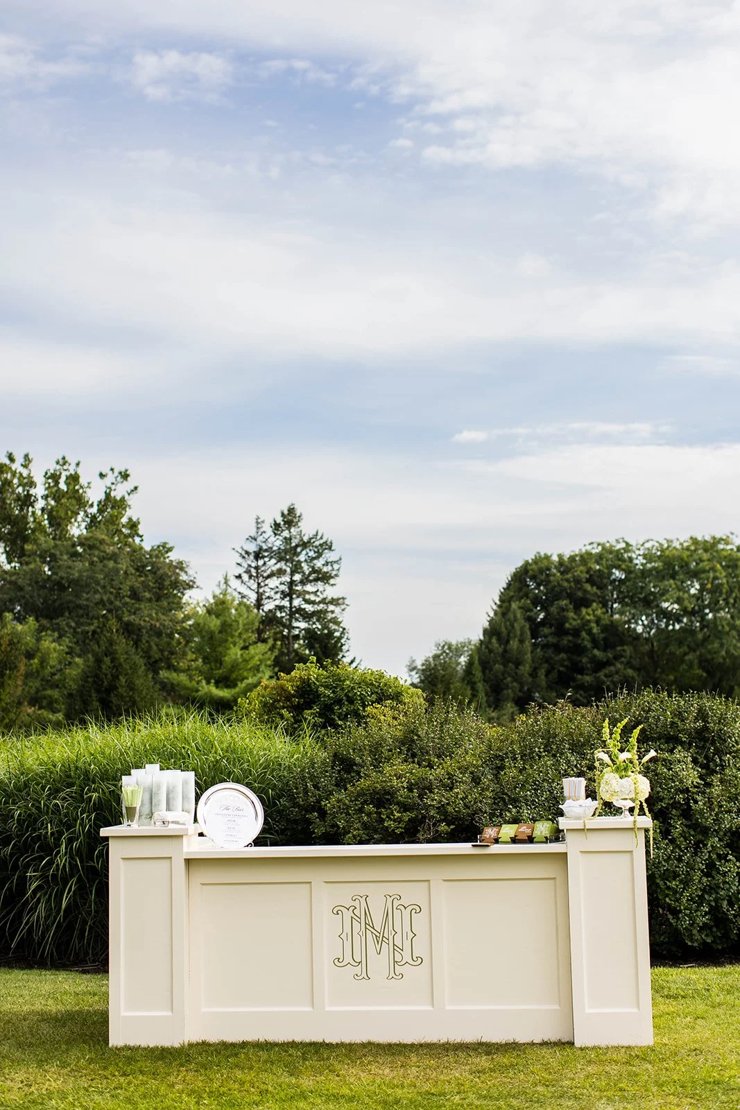 White outdoor bar with monogram 'M' on front panel, decorated with flowers and event signage, set against greenery and blue sky.