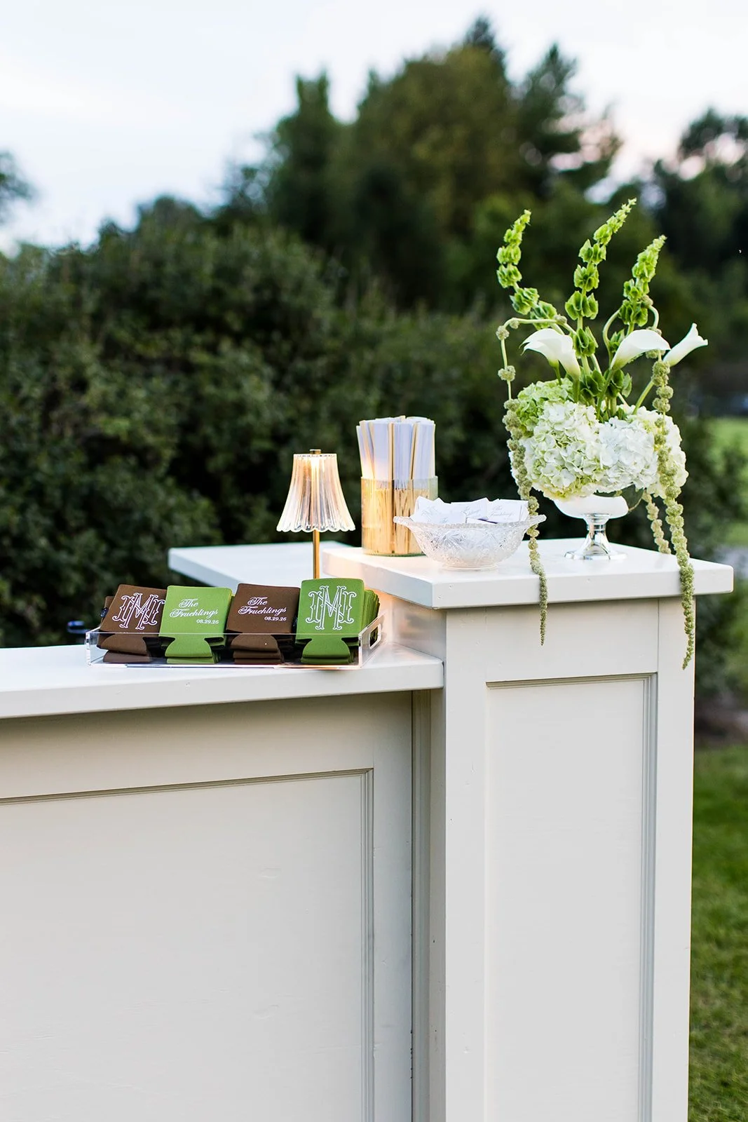 White reception table outdoors with green and brown tags labeled 'Mr' and 'The Furchungs', a small table lamp, a vase with white flowers and greenery, and a bowl with papers, set against a background of trees and sky.