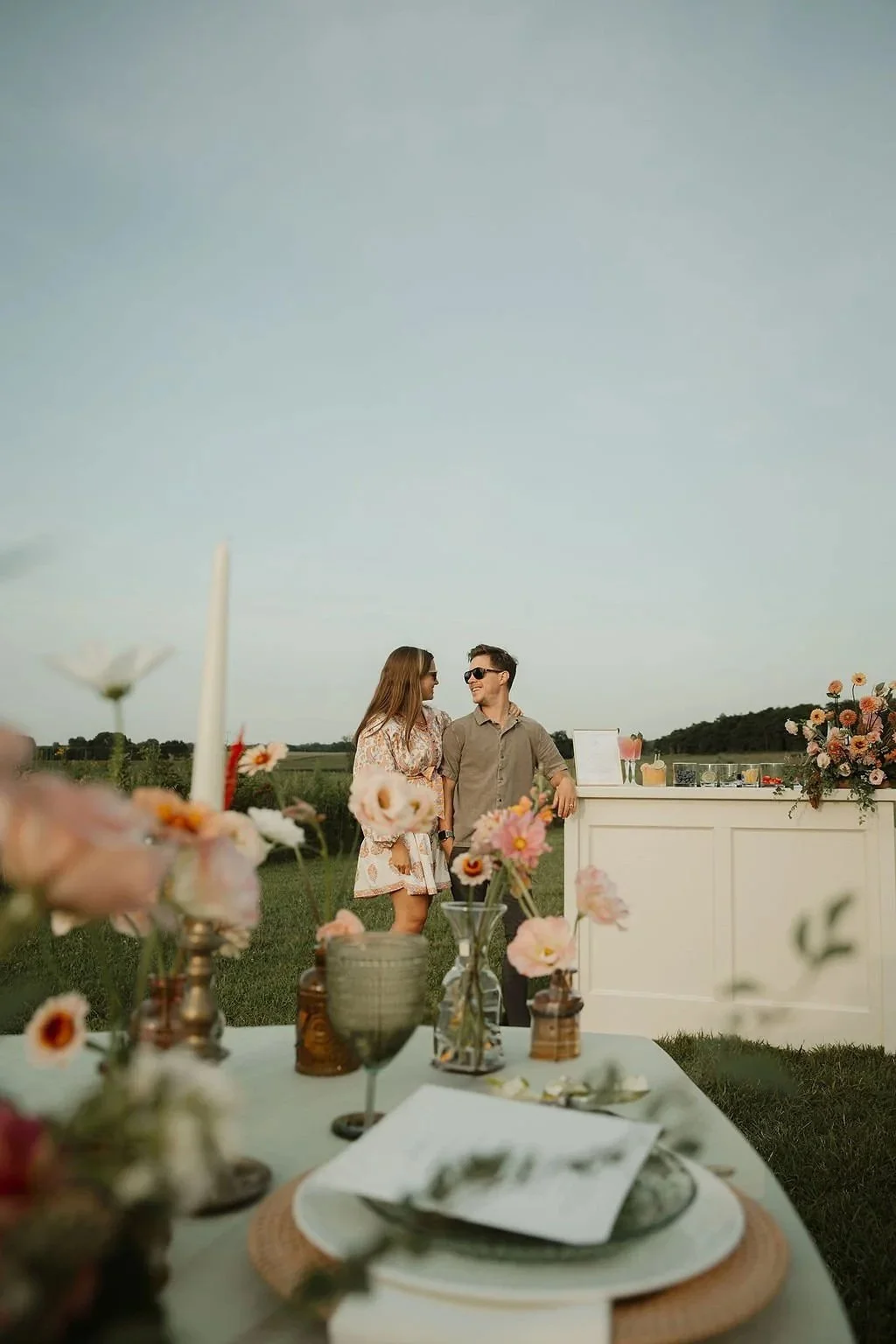 A couple standing outside at a garden party, with a table of flowers and food in the foreground. The couple is smiling at each other near a white bar or counter, with a landscape and clear sky in the background.