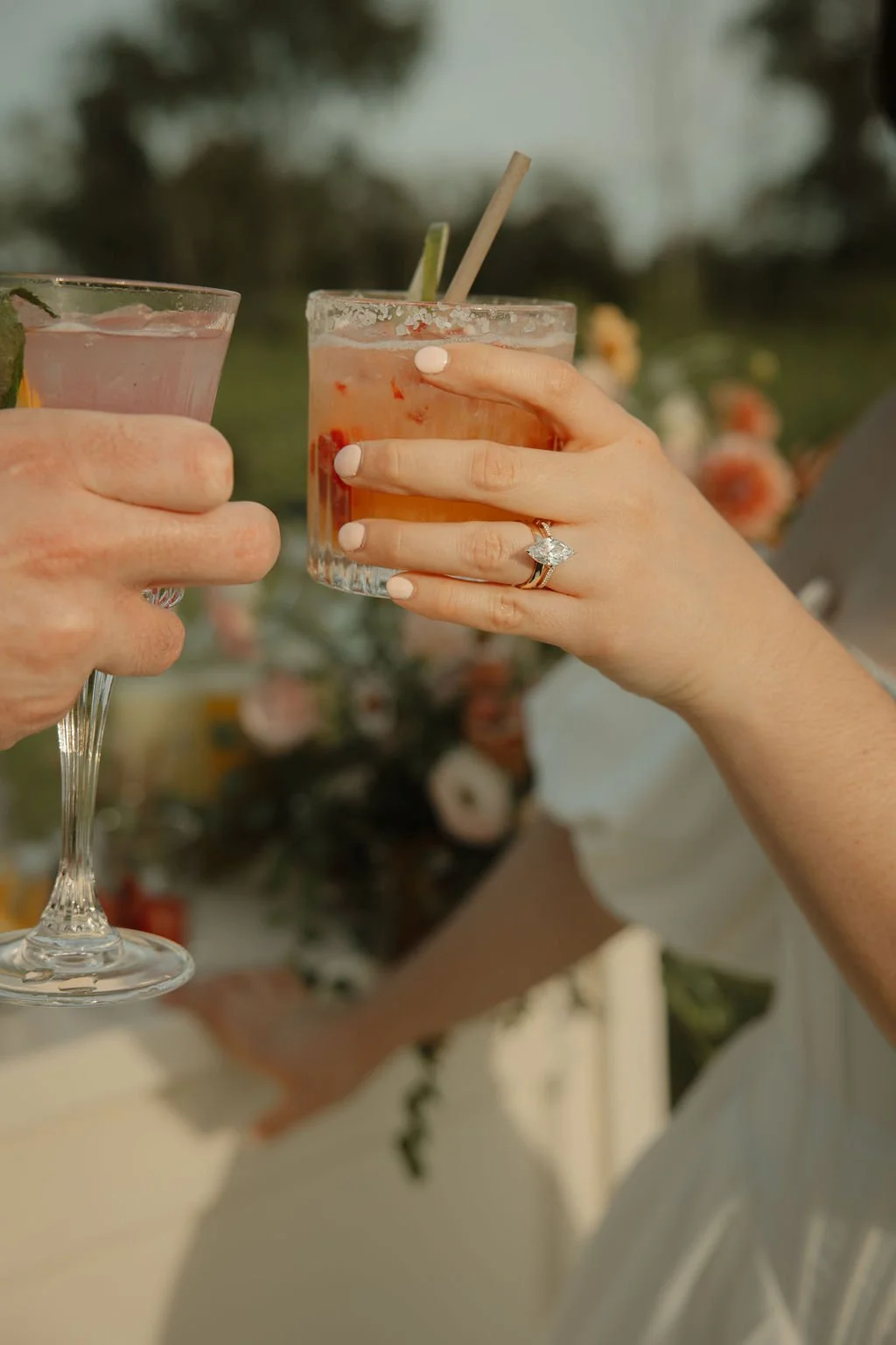 Two people clinking glasses, one with a cocktail and the other with a sparkling beverage. One person is wearing a large diamond ring.