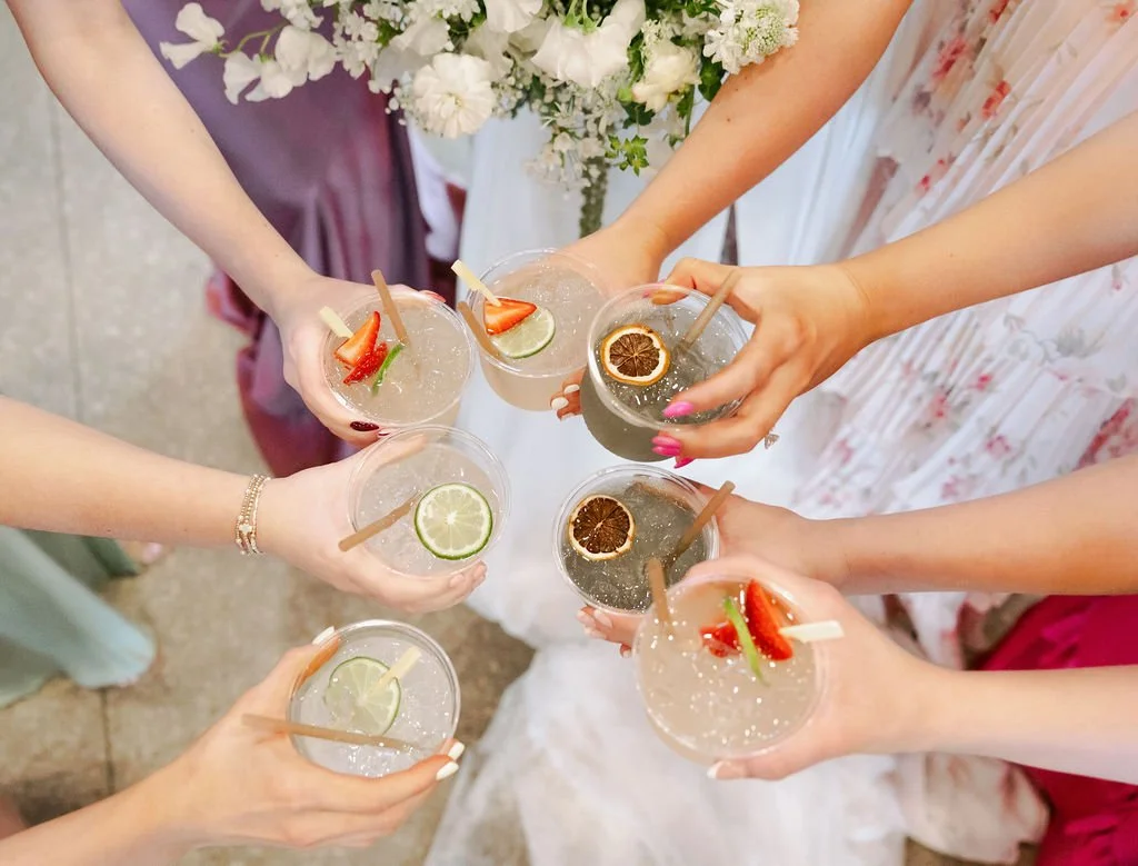 Several women holding cocktails with fruit garnishes, including lime, strawberry, and dried citrus slices, at a celebration or gathering.