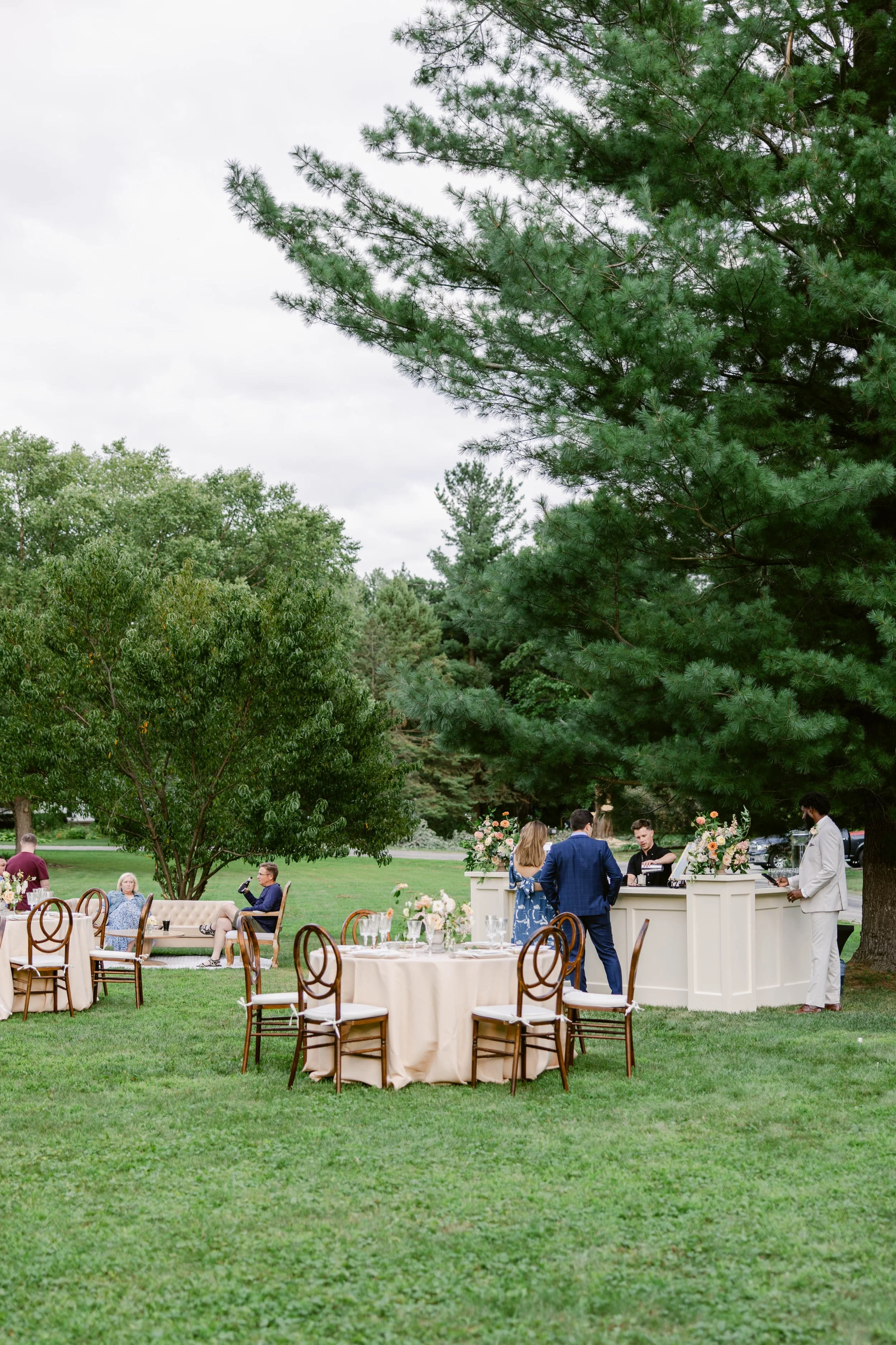 An outdoor wedding reception with round tables covered in beige tablecloths and decorated with flower centerpieces. Guests are seated and socializing on a lush, green lawn surrounded by large trees. A white bar or serving counter is set up where peop