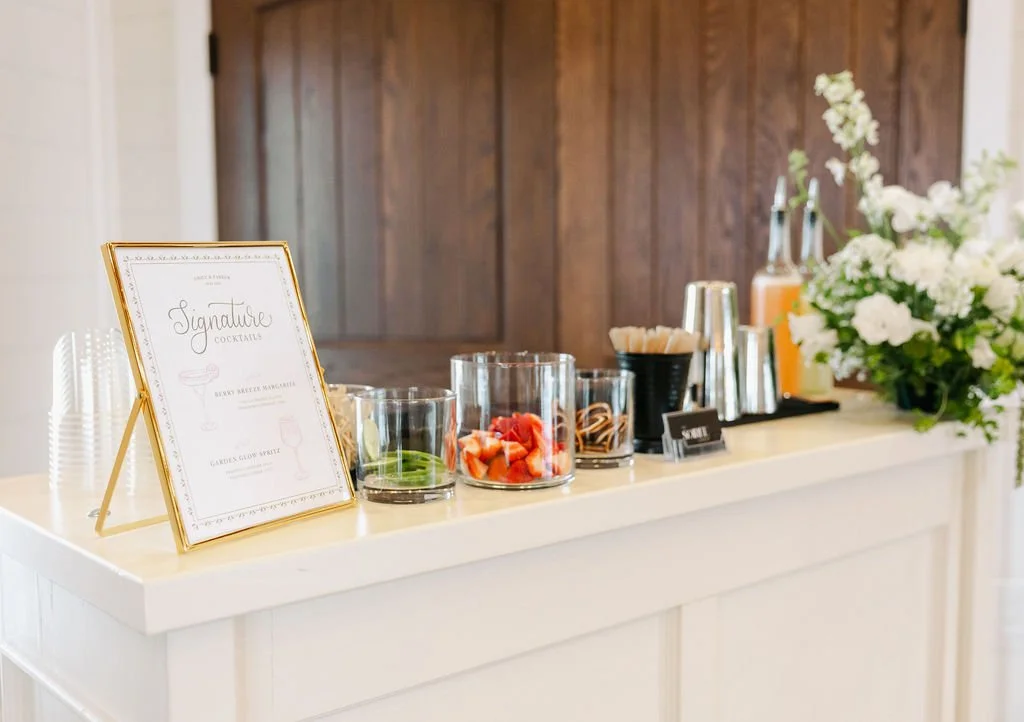 A white bar or table with a sign reading 'Signature Cocktails' and various drink ingredients in glass jars, along with plastic cups, stirrers, and a large floral arrangement with white flowers and greenery.