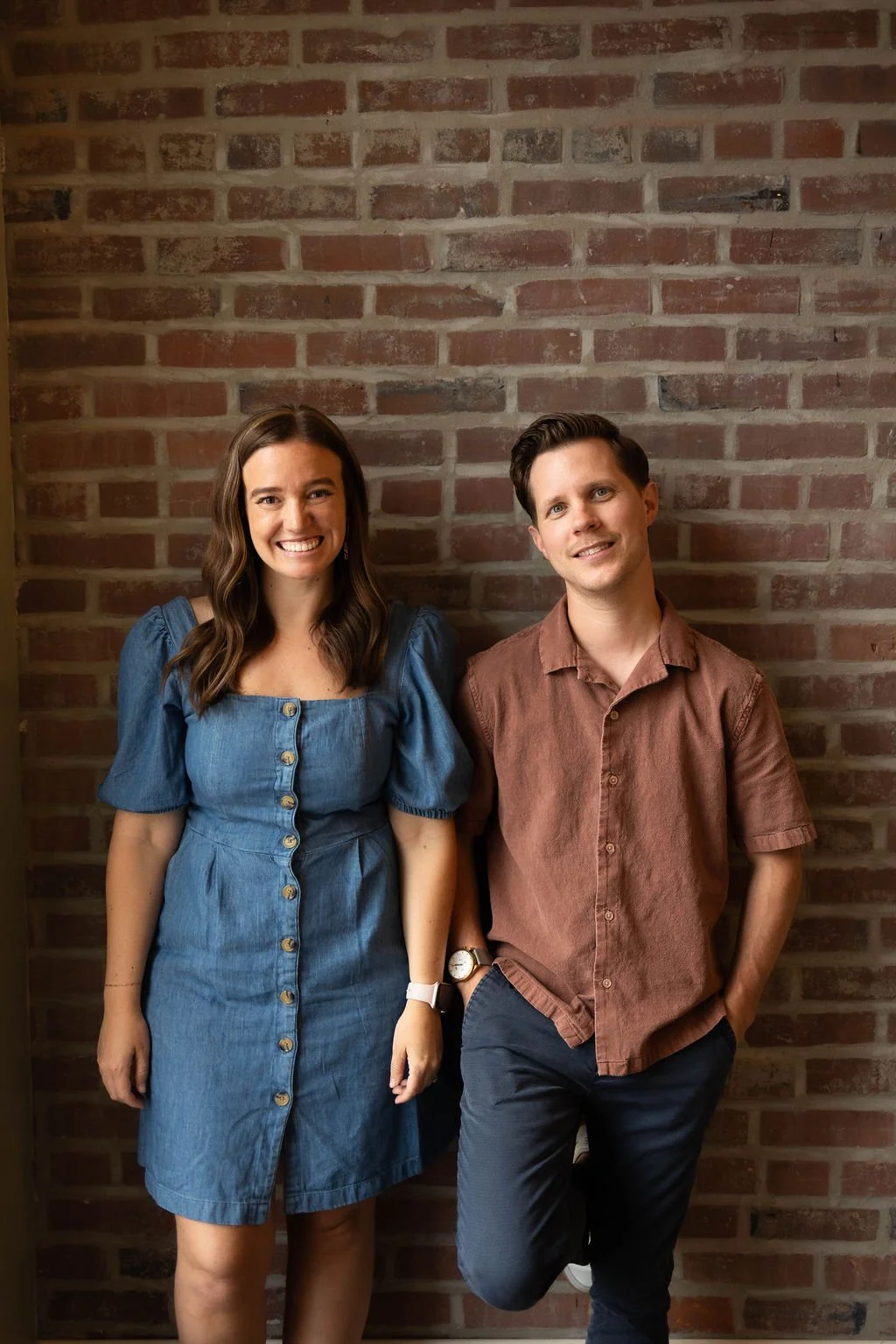 A smiling woman with long brown hair in a blue denim dress with buttons, standing next to a smiling man with short brown hair in a brown short-sleeved shirt and dark pants, against a brick wall background.