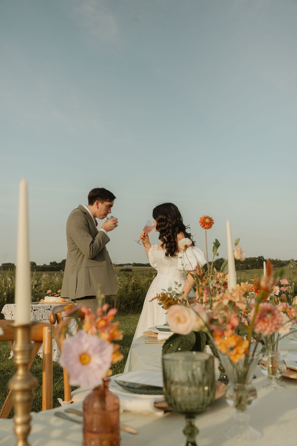 A man and woman in wedding attire having drinks during an outdoor wedding reception with decorated tables and floral arrangements.