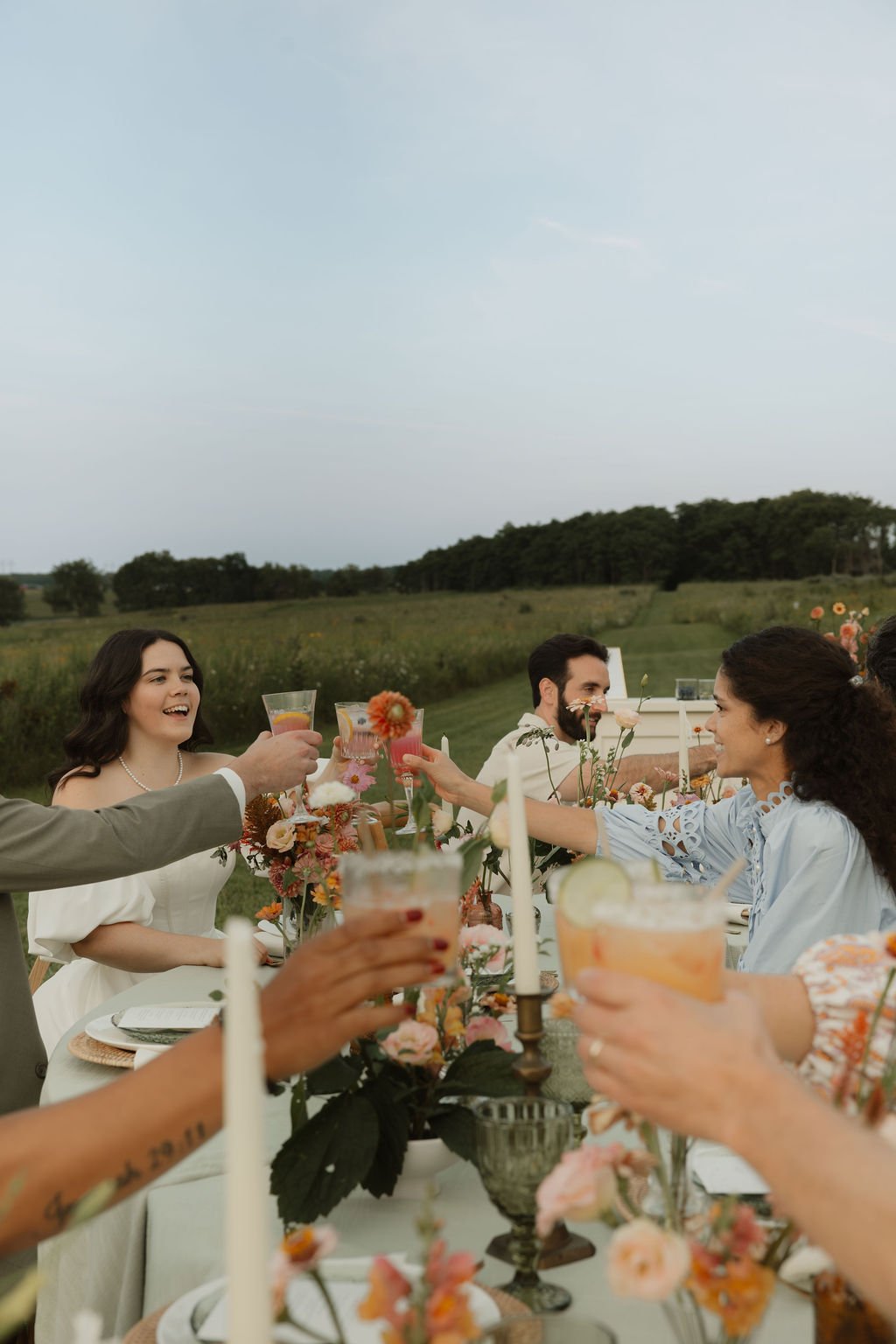 People raising drinks at an outdoor celebration table with floral decorations, set against a green field and cloudy sky.