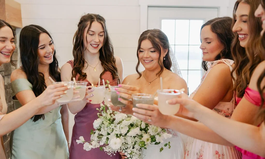 Group of women at a wedding celebration, clinking drinks and smiling, with a bride holding a bouquet.