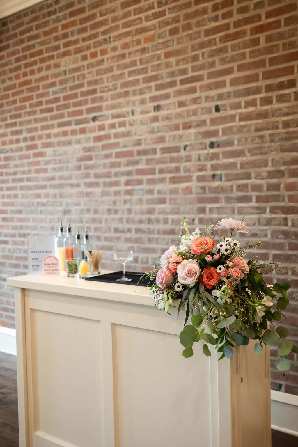 A floral arrangement with pink, white, and peach roses, white daisies, and greenery on a cream-colored bar against an exposed brick wall. In the background, there are bottles, small containers, and a sign on the bar.