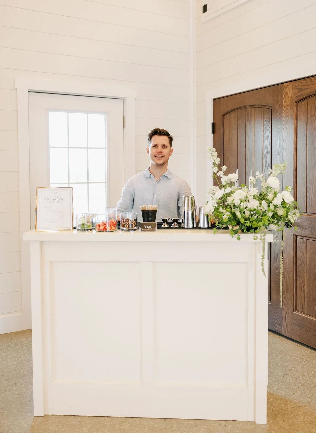 A smiling man standing behind a white bar or counter with glass jars, a framed sign, and a large floral arrangement, inside a bright room with white walls and wooden doors.
