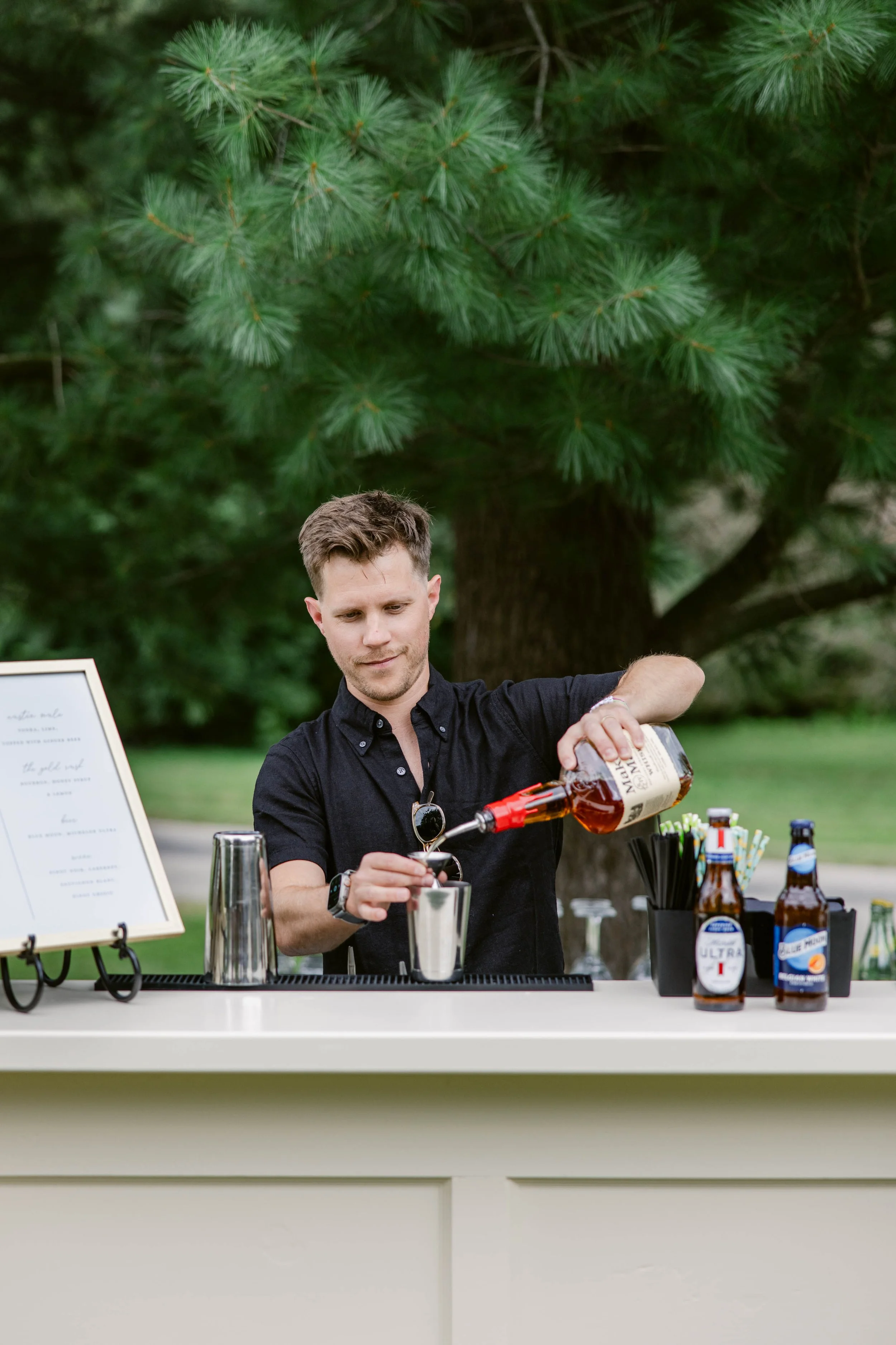 A young man in a black shirt pouring a drink into a glass at a bar outdoors with trees in the background. Bottles of beer and cocktail mixers are on the bar counter.