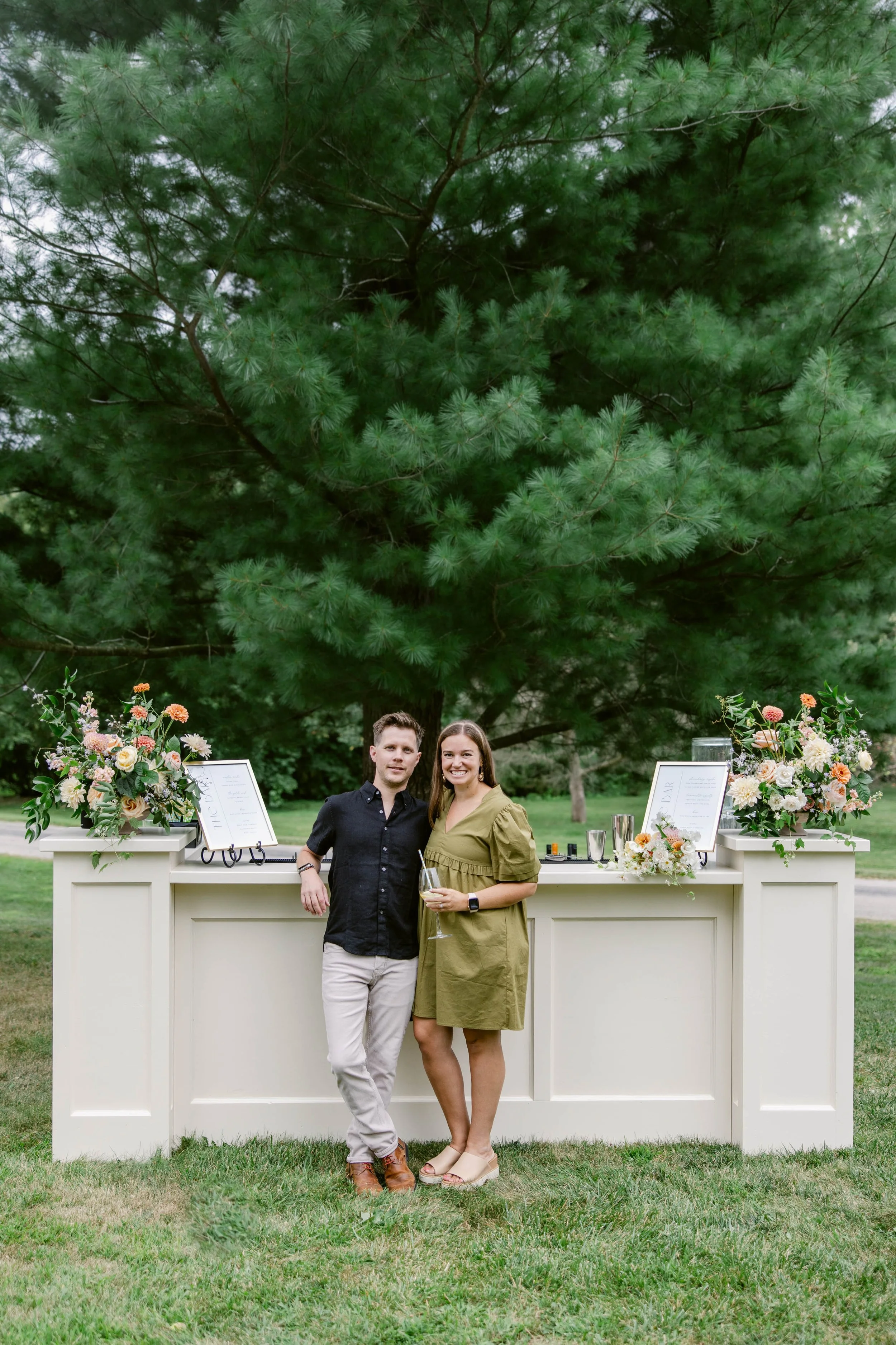 A couple standing outdoors in front of a white bar with floral arrangements, wine glasses, and framed menu inserts, under a large green pine tree, holding glasses of wine and smiling at the camera.