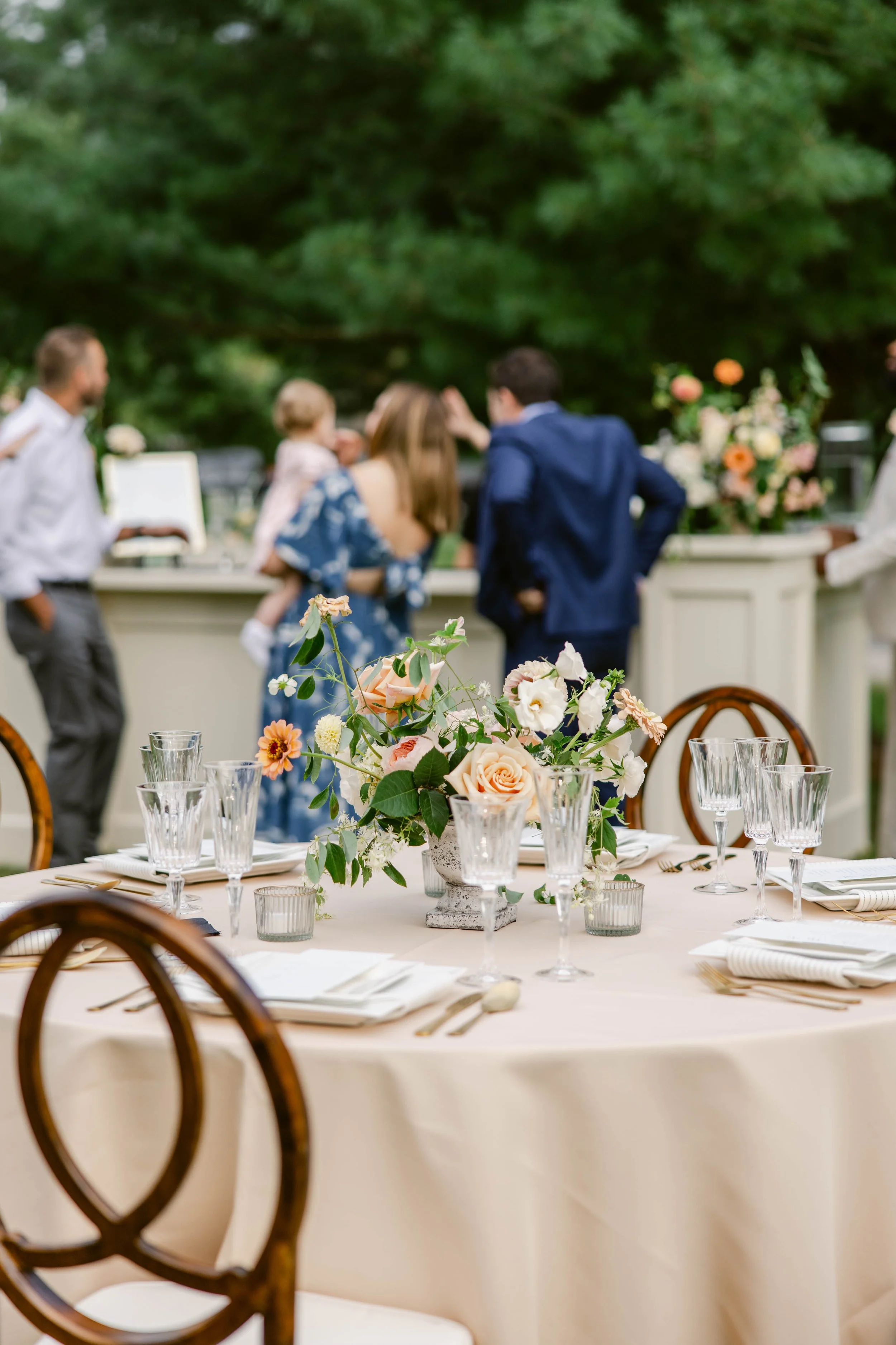 A round table with a cream tablecloth, decorated with a floral centerpiece and crystal glassware, set for a formal event; blurred background shows guests talking and a lush, green outdoor setting with trees and flowers.