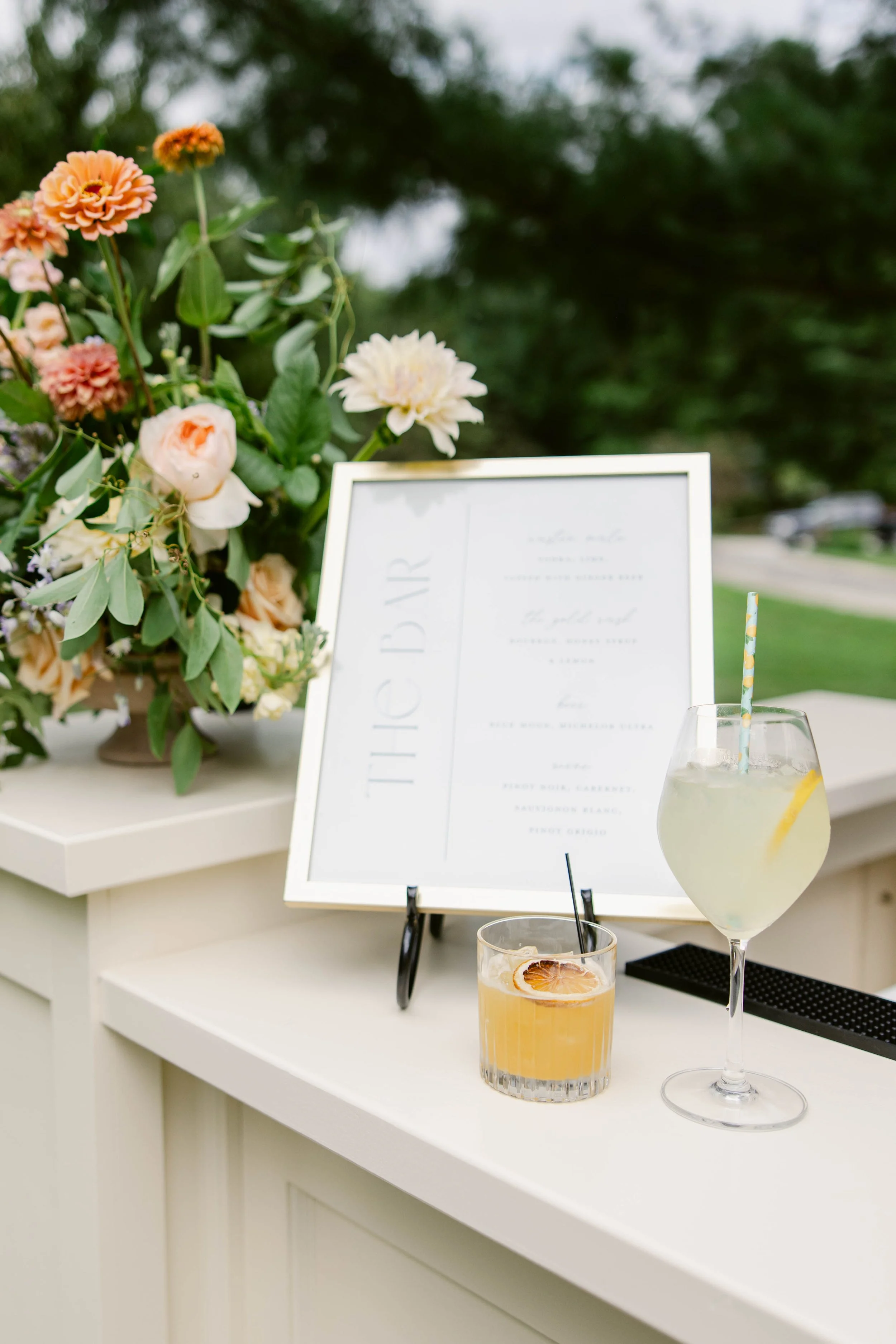 A floral arrangement on a white table with two glasses of drinks, a wine glass with a yellow cocktail and a smaller glass with an orange drink and a lemon slice. A framed menu is in the background with blurred text.