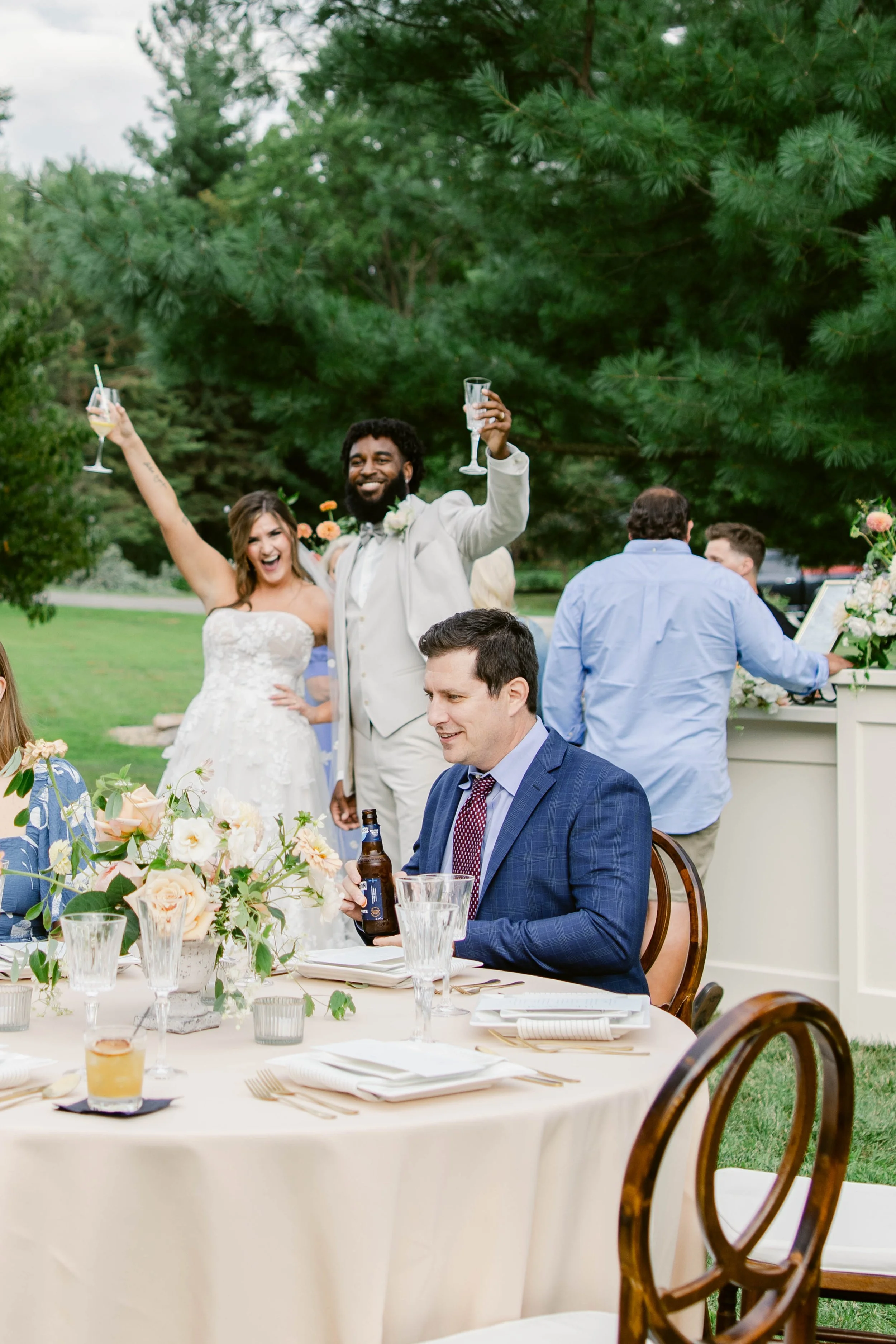 People celebrating outdoors, with one couple raising glasses, surrounded by greenery, table with floral centerpiece, and guests in formal attire.
