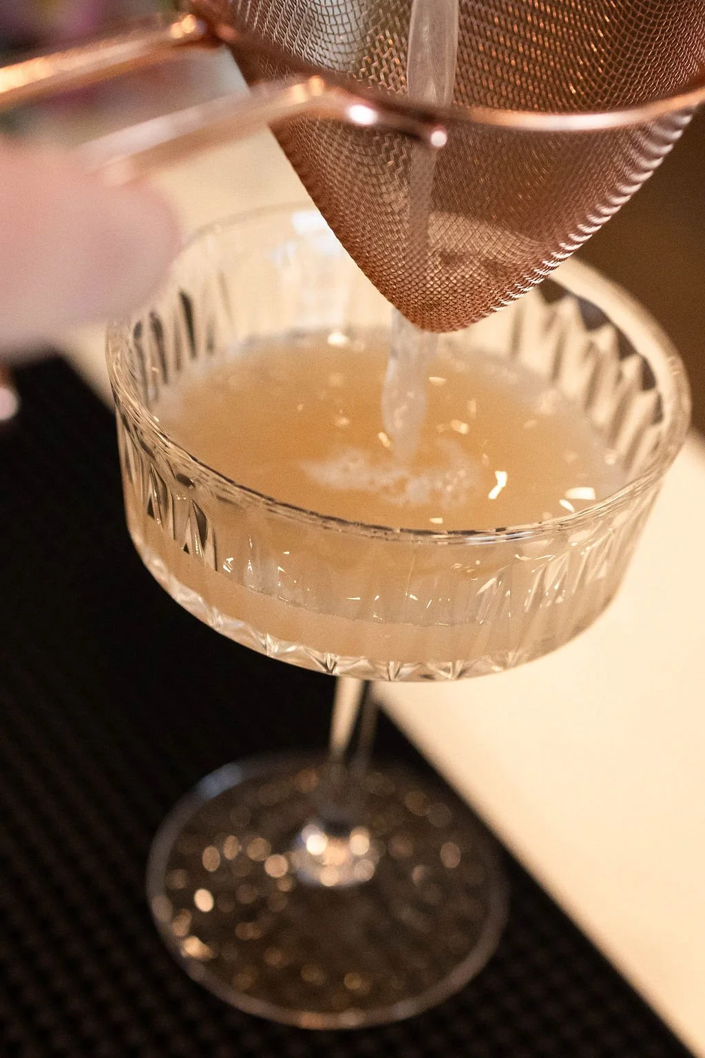 A mesh strainer filtering a liquid into a crystal glass, which is placed on a dark surface.