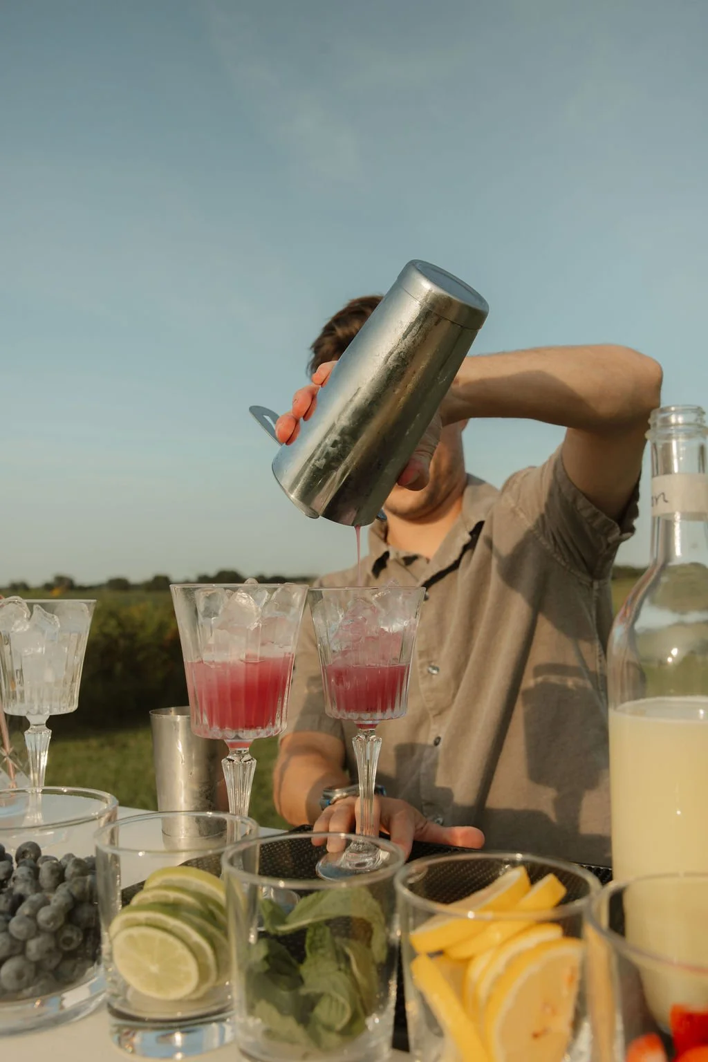 A person pouring a pink beverage into glassware on an outdoor table with fruit garnishes and a bottle of yellow liquid.