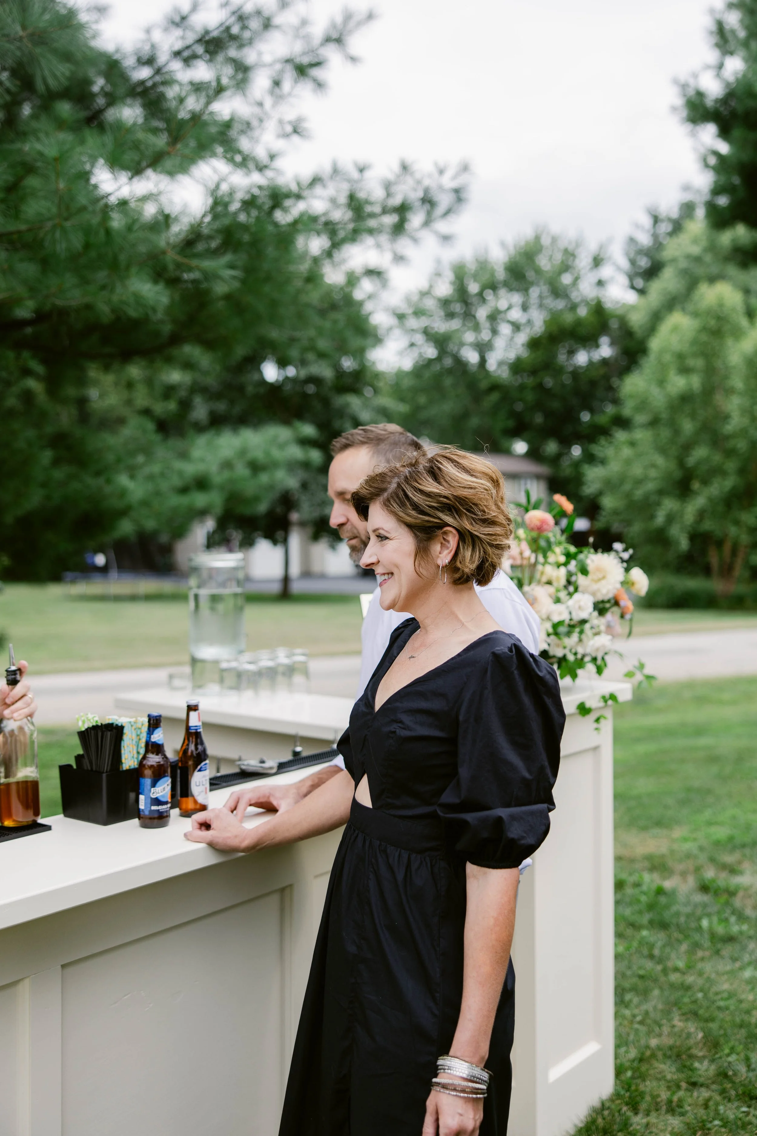 A woman with short, wavy brown hair and silver bracelets, wearing a black dress, standing at an outdoor bar with a man behind her. The bar has bottles of alcohol and drink garnishes, with trees and houses in the background.