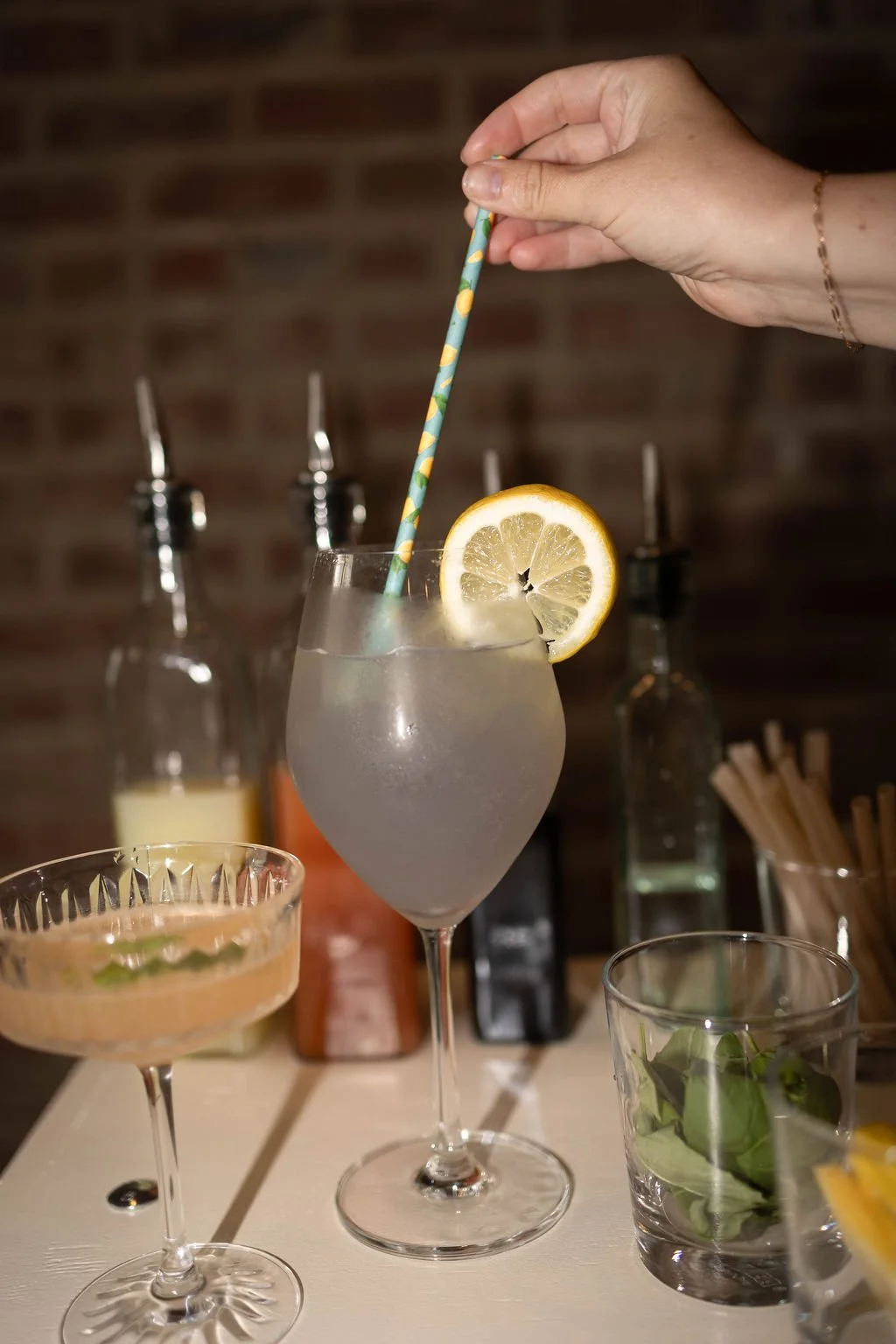 A hand holding a colorful straw in a clear glass garnished with a lemon slice, with various cocktail ingredients and glasses on a bar backdrop.