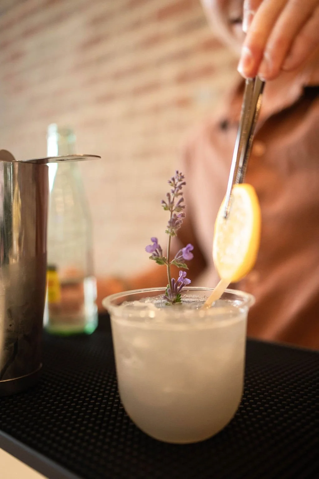 A person preparing a cocktail with a lemon slice and a sprig of lavender garnishing the drink.