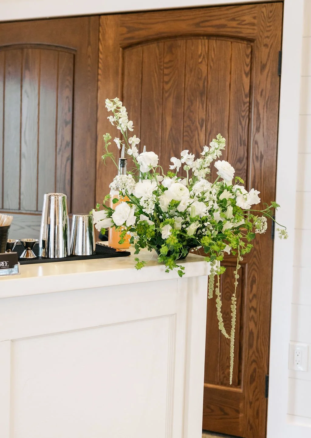White floral arrangement on a bar counter with stainless steel cups and bar tools, set against wooden cabinets in a kitchen.