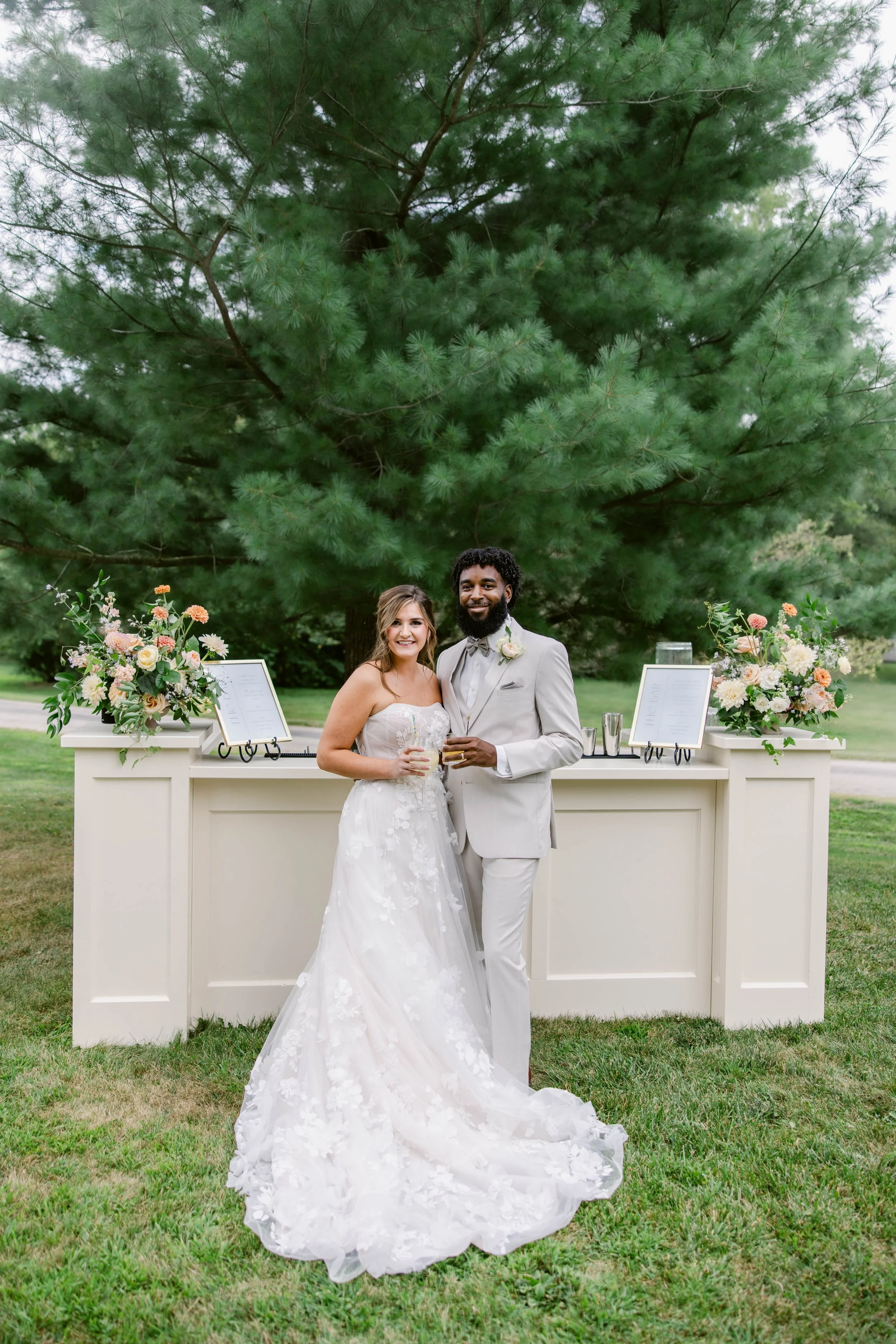 A newlywed couple standing outdoors next to a white bar with floral arrangements, with a large green pine tree in the background, smiling and holding drinks.
