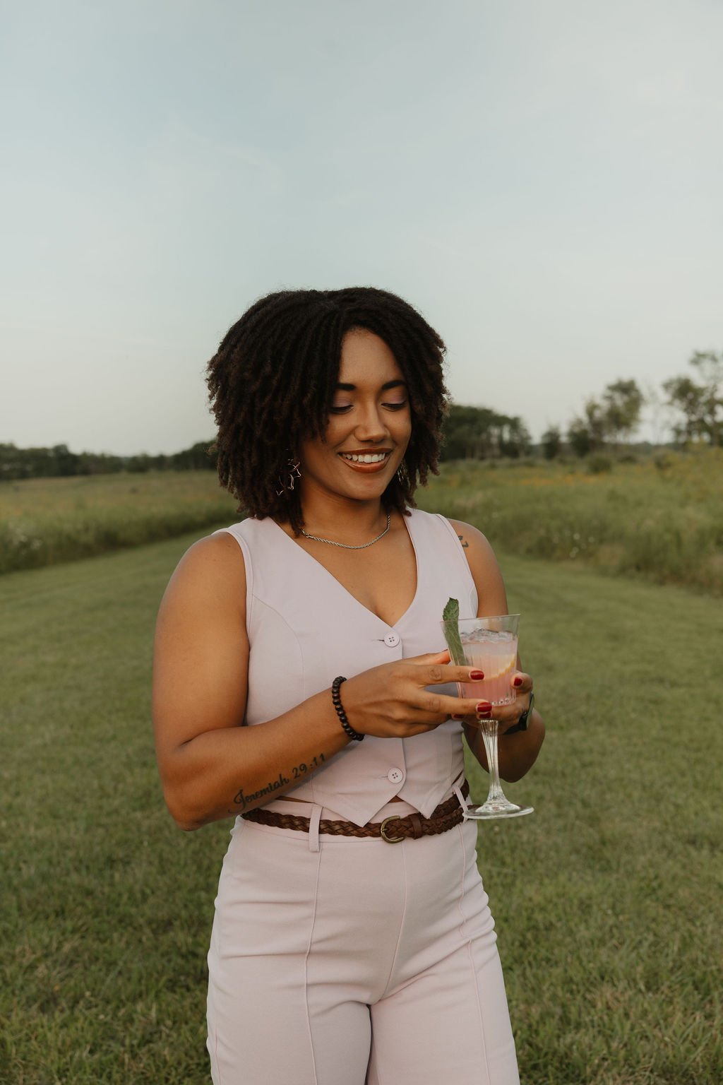 A woman with dark curly hair, wearing a light-colored sleeveless top and pants, is holding a cocktail glass with a green garnish, standing outdoors in a grassy field during daytime.
