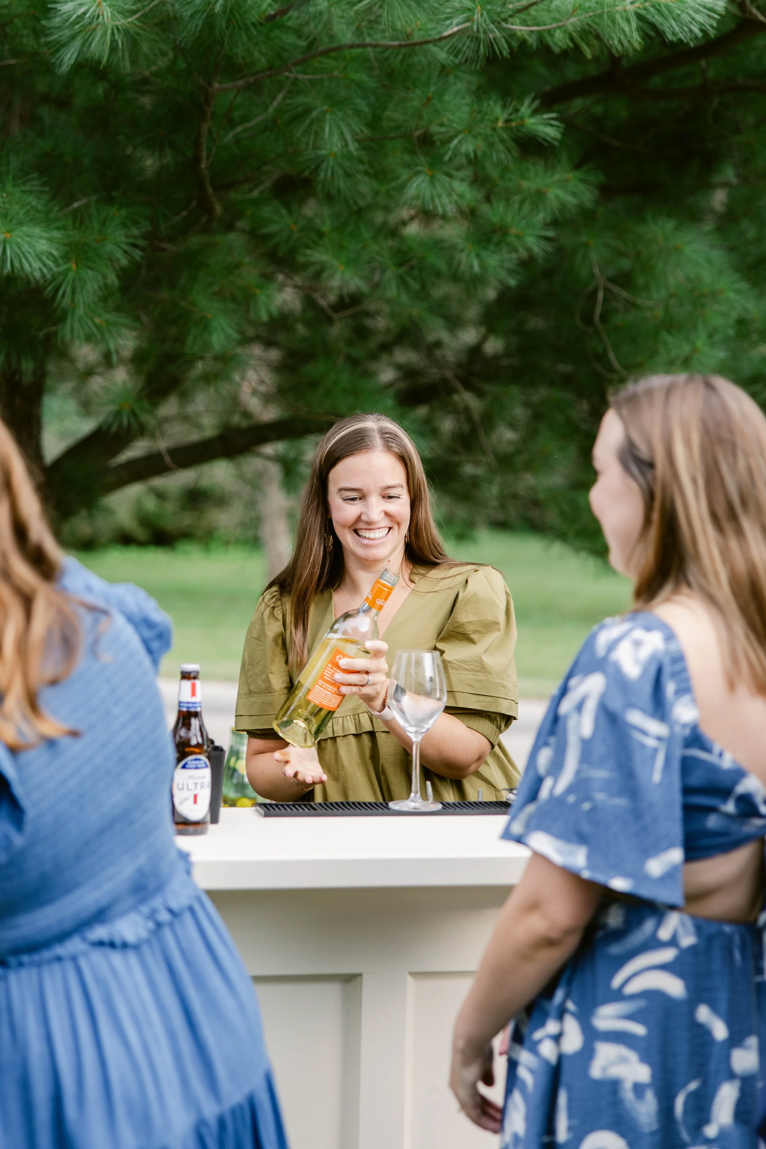 A woman with long brown hair smiling as she pours wine into a glass at an outdoor gathering with other women, under a tree with green pine needles. The group is around a table with bottles and glasses, enjoying a social moment.