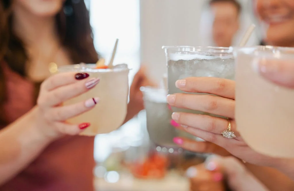 People clinking glasses with drinks, including a pink frozen cocktail with an umbrella and a clear beverage, during a social gathering.