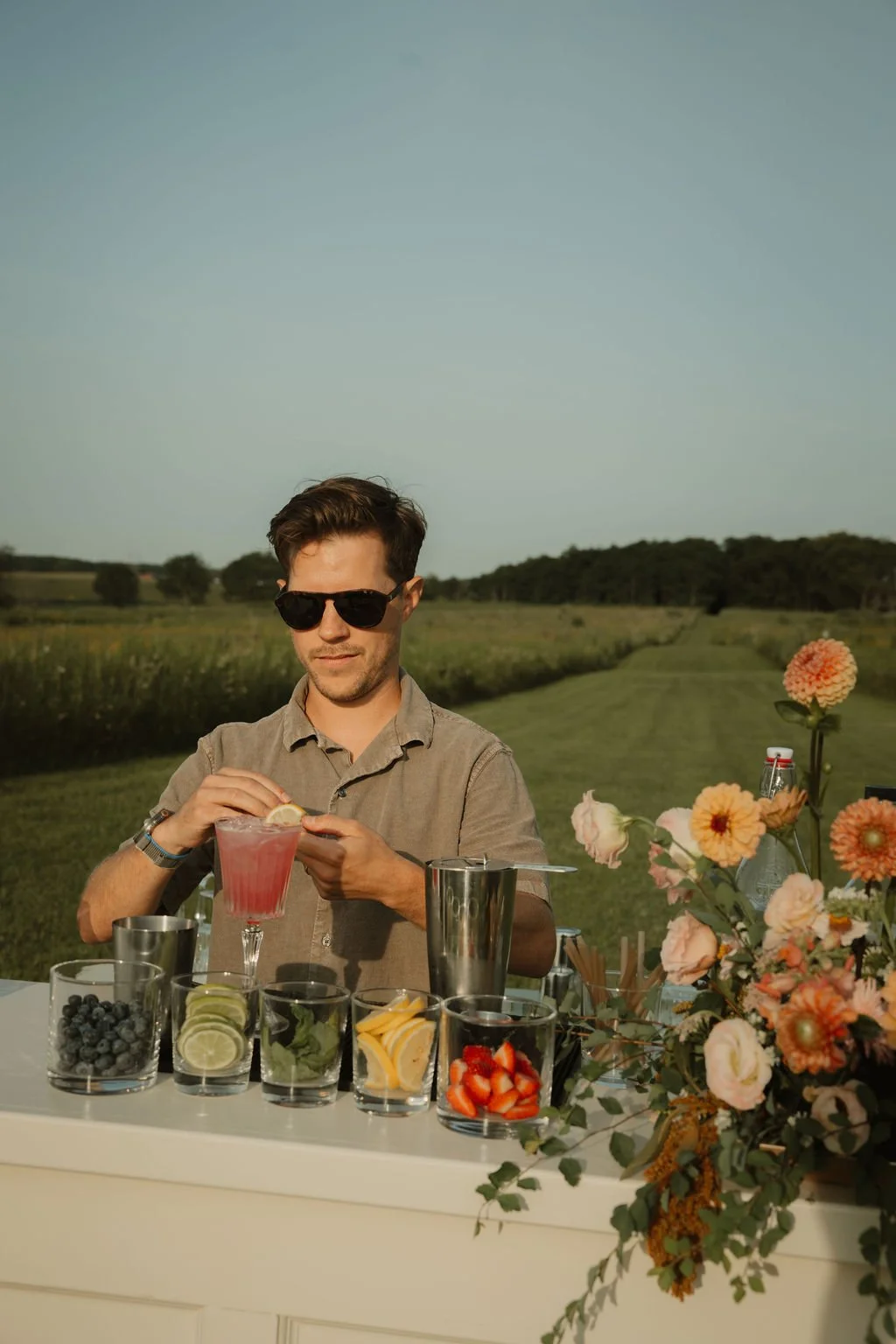 A man wearing sunglasses and a beige shirt is preparing a pink cocktail with lemon slices at an outdoor table set on a grassy field with a clear sky and distant trees. The table has various fruits, a metal shaker, and a flower arrangement.