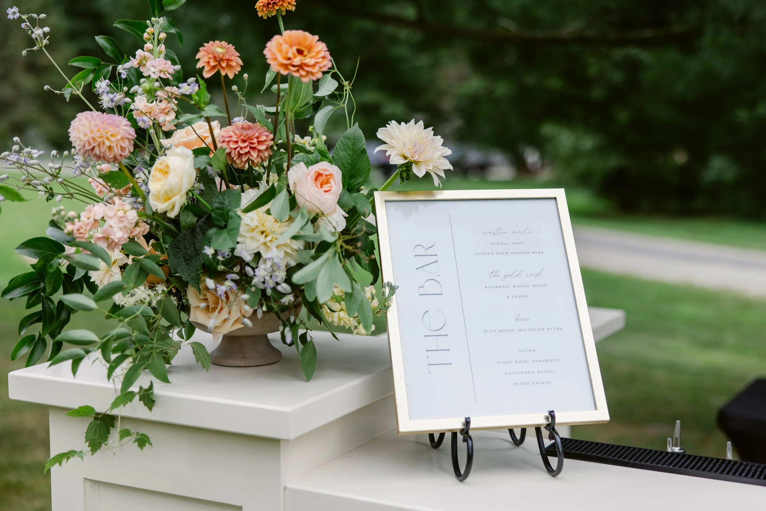 A floral arrangement with pink, white, and peach flowers on a table at an outdoor event, with a white sign displaying drink options.
