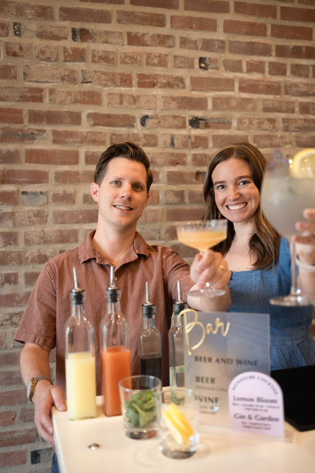 A man and woman smiling and holding up drinks, standing behind a bar with bottles of various beverages, a lemon garnish, and a menu with options for beer, wine, and cocktails, against a brick wall background.