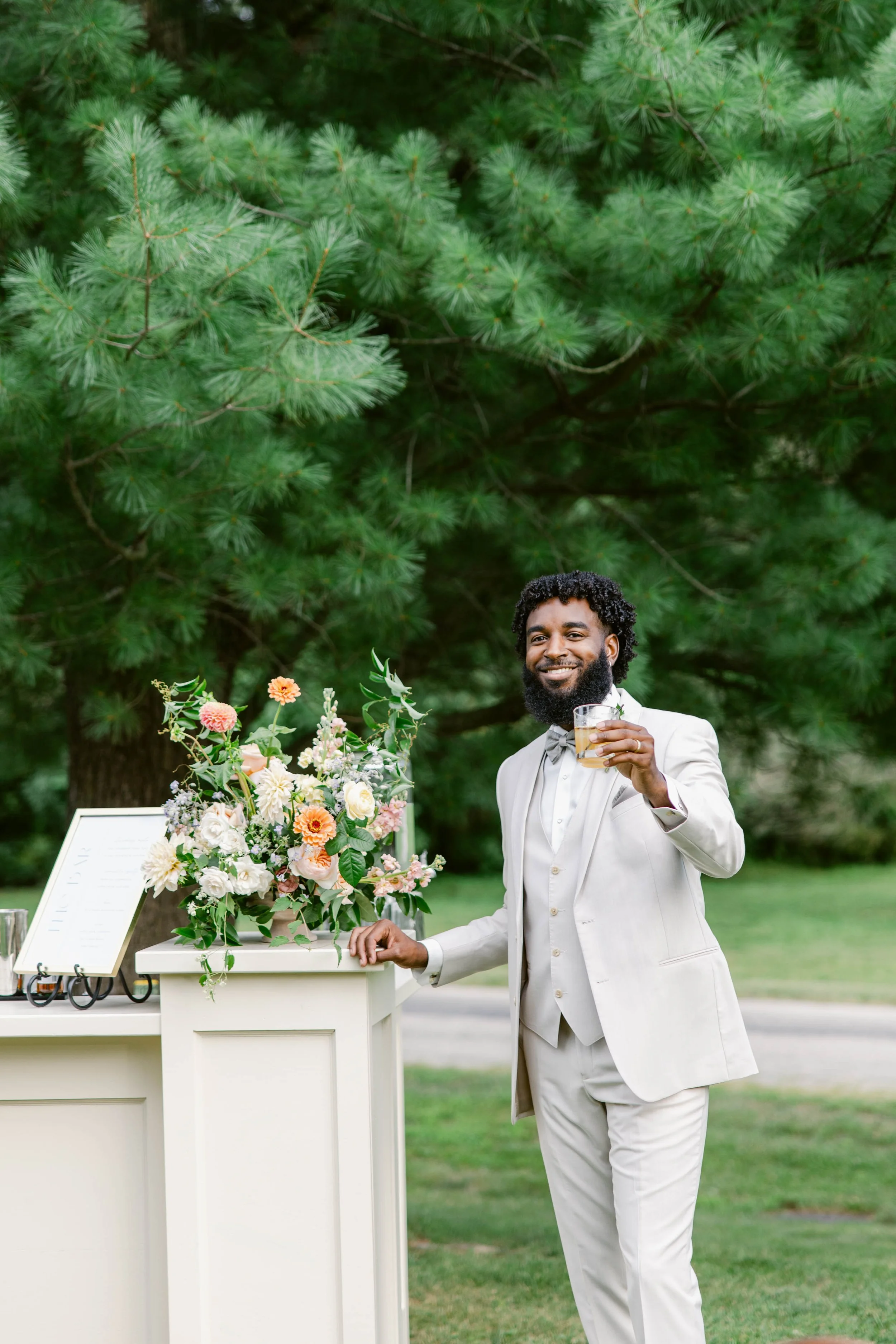 A man in a white tuxedo smiling and holding a glass of champagne standing outdoors near a floral display and a white table, with green trees in the background.