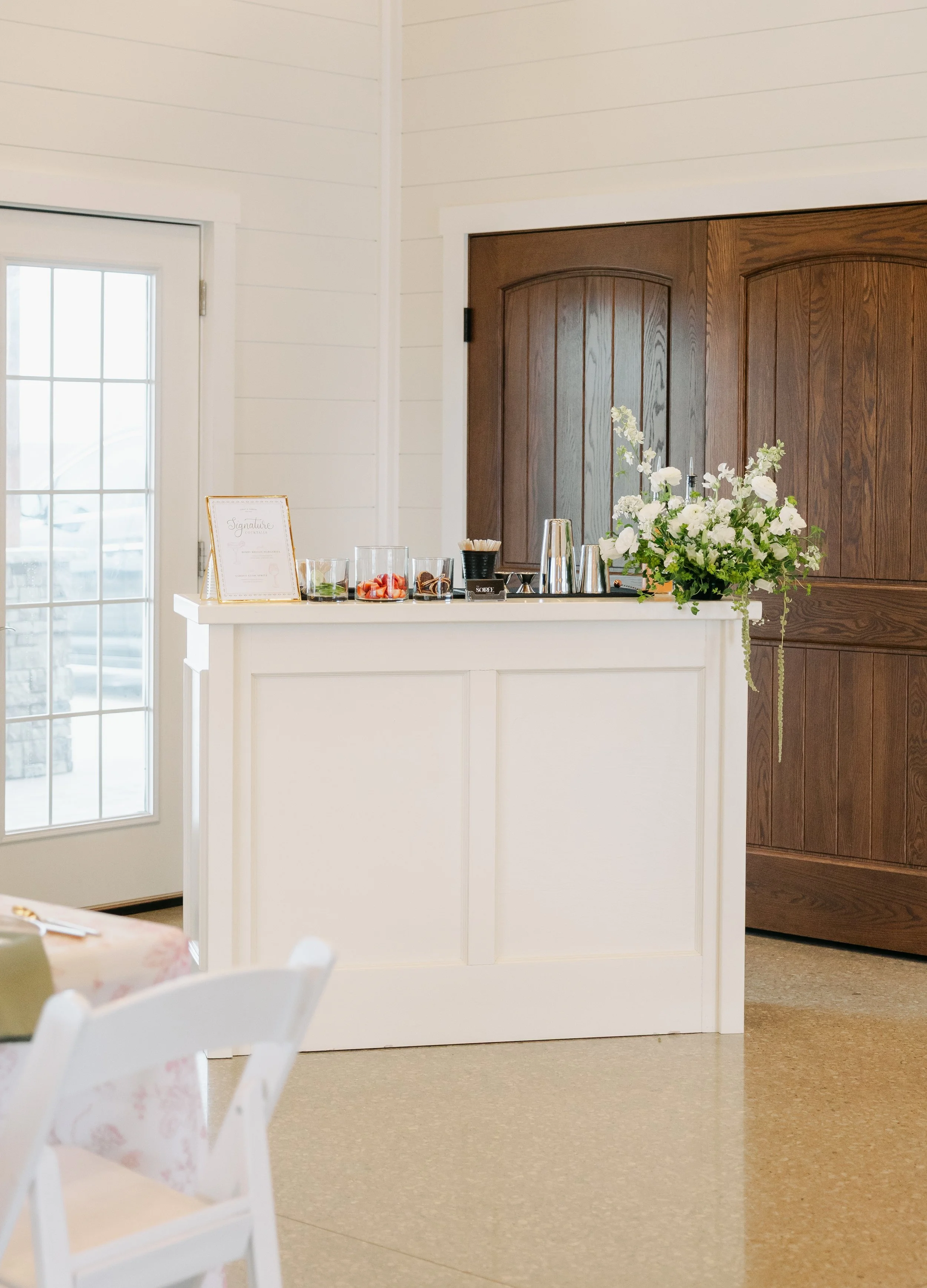 A white counter with a floral arrangement, fruit, and drink station in a room with cream-colored walls and wood accents, possibly for an event.