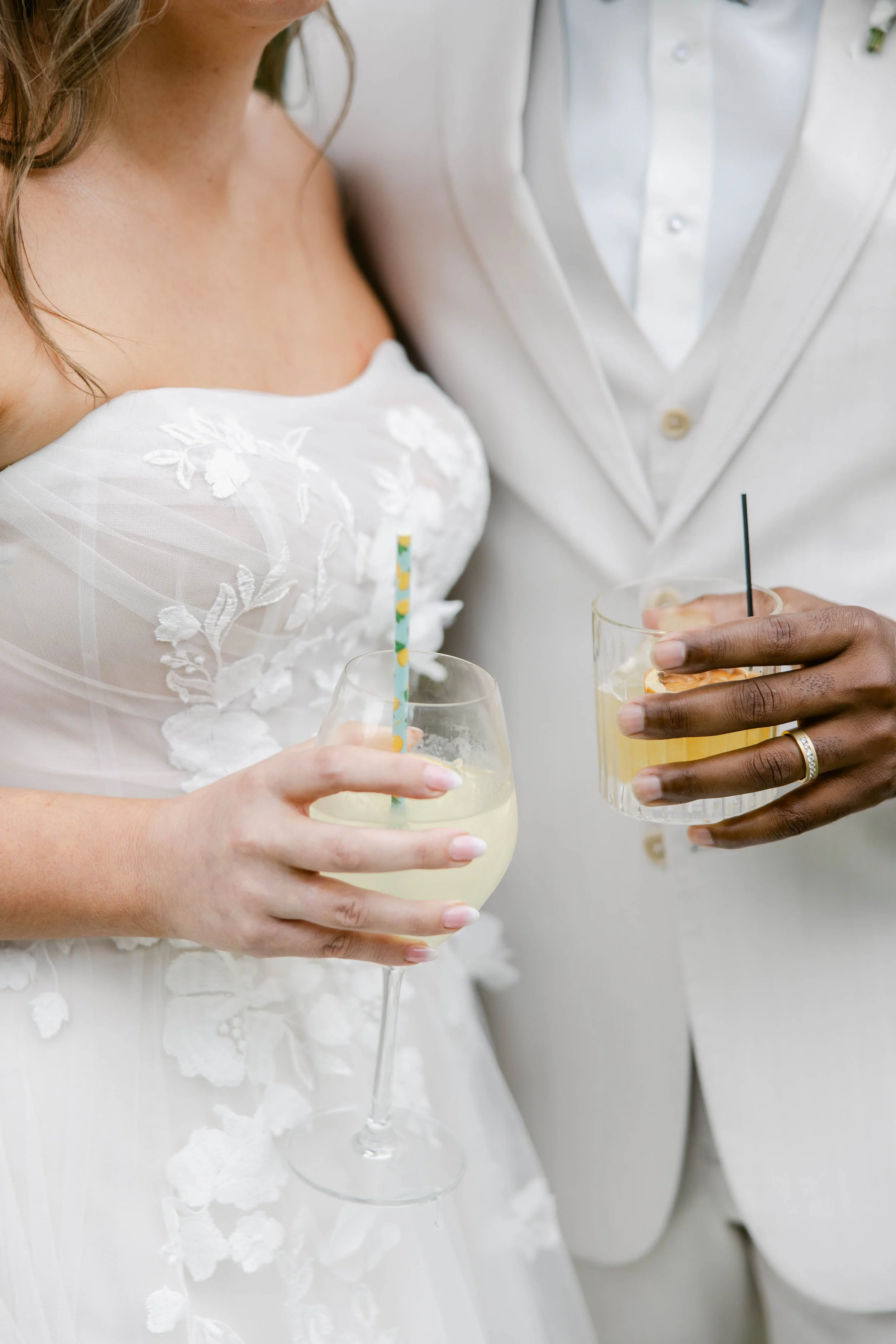 Close-up of a couple at a wedding, holding wine glasses with drinks and straws. The woman is wearing a white dress with floral embroidery, and the man is dressed in a white tuxedo with a shirt and bow tie.