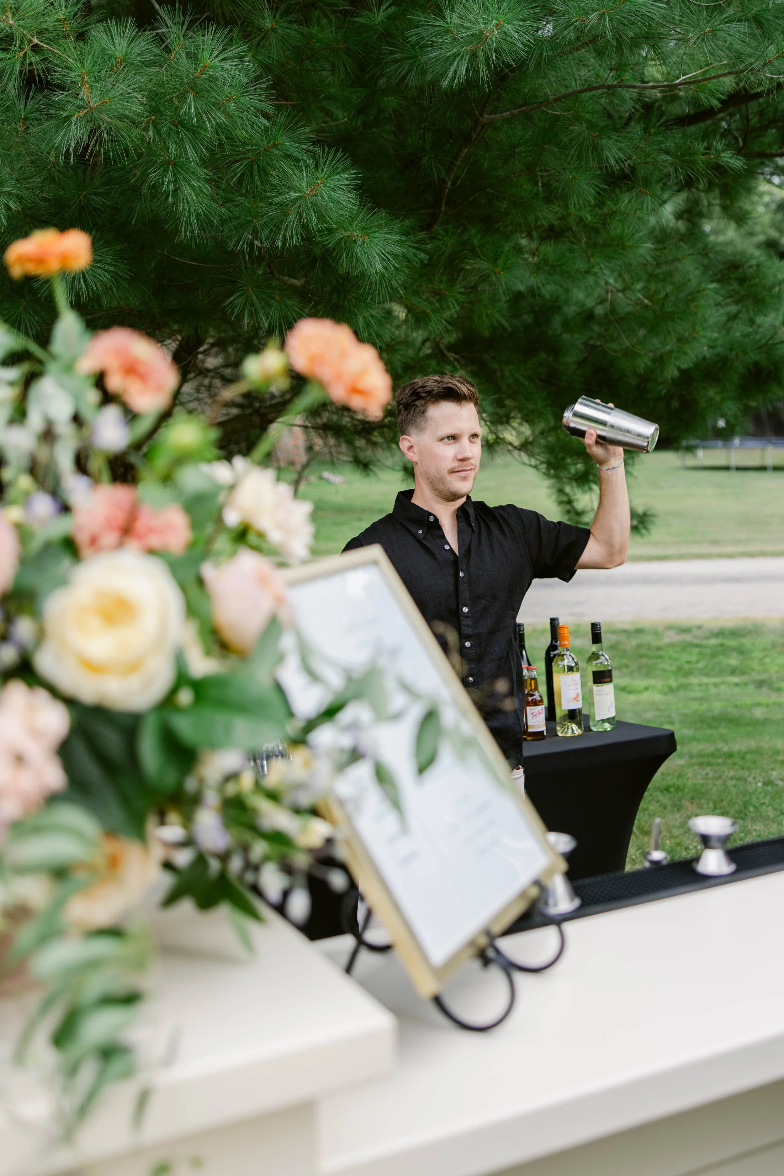 A man standing outdoors under pine trees, holding a metal cocktail shaker. In the foreground, blurred flowers and a display of bottles and drinks are visible.