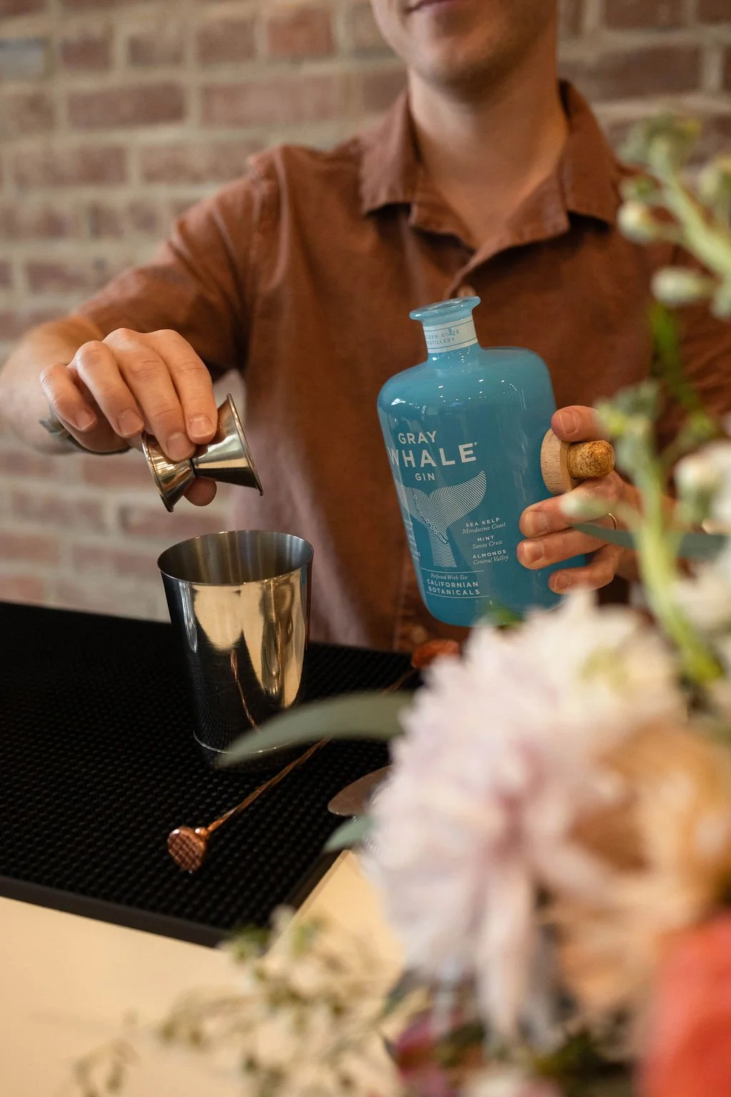 A person in a brown shirt pouring gin from a blue bottle marked 'Gray Whale Gin' into a metal jigger over a cocktail shaker, with a brick wall in the background and some flowers in the foreground.