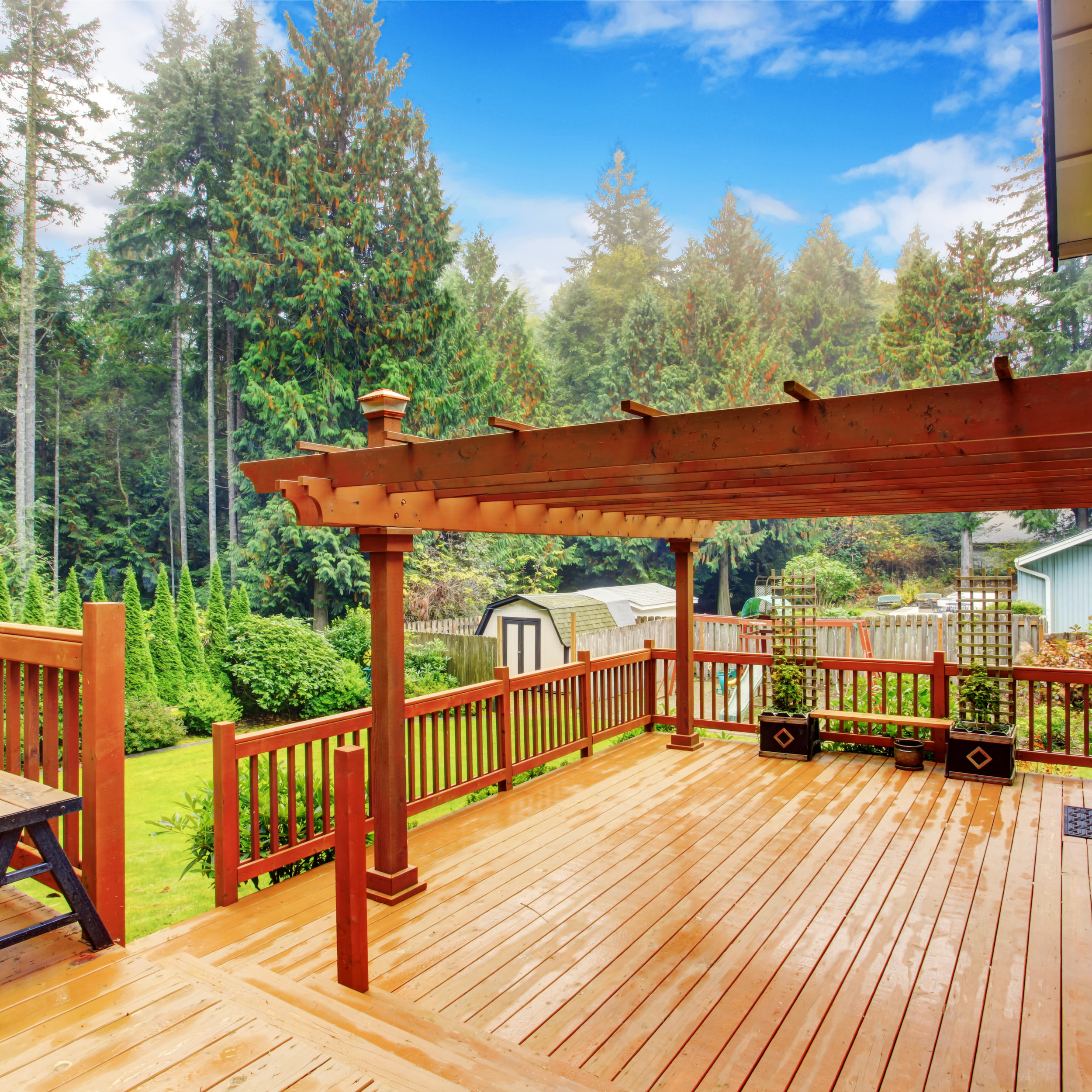 Wooden deck with a railing, overlooking a lush green backyard with trees and shrubs, under a partly cloudy blue sky.