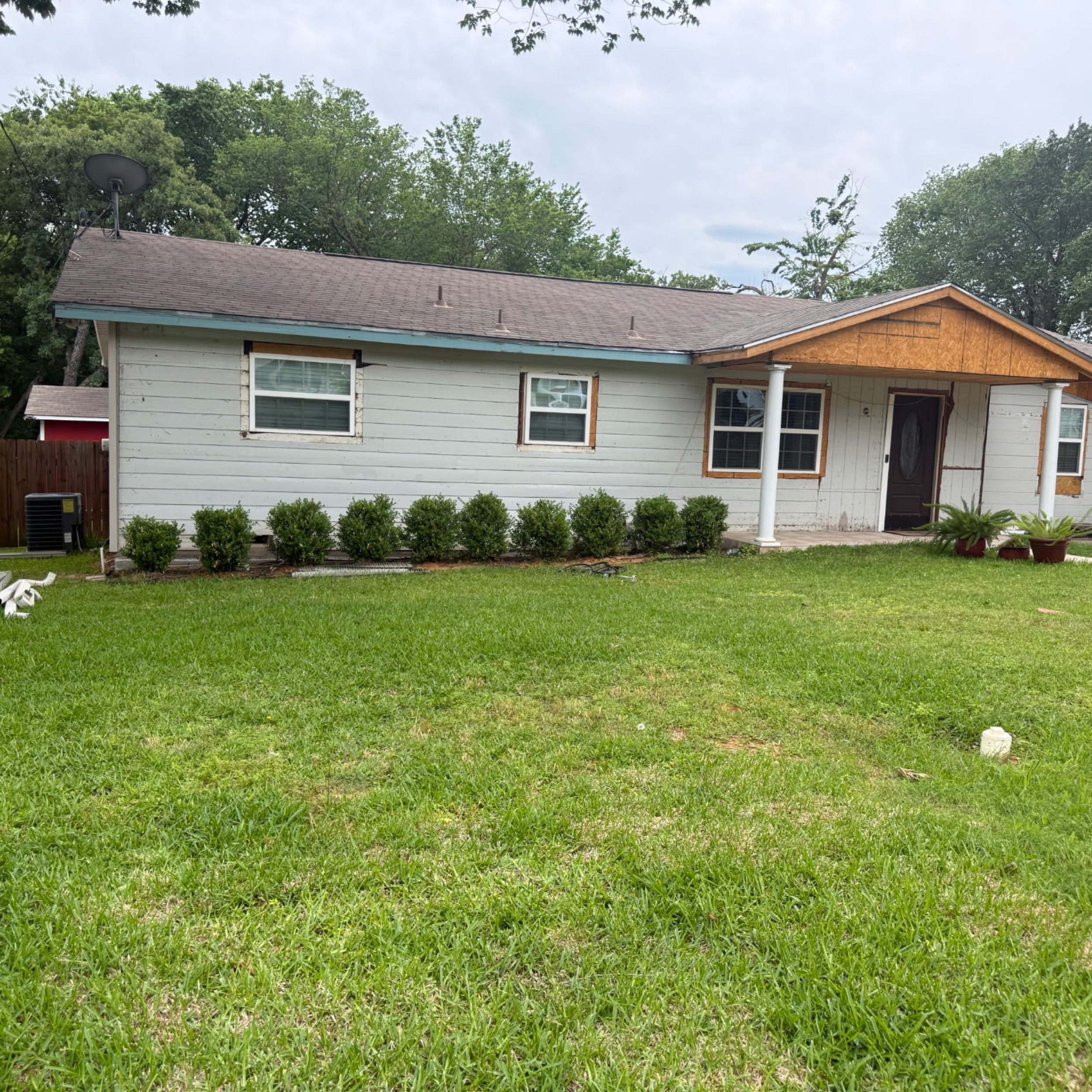 View of a house with a brown roof, white exterior walls, multiple windows, a front porch supported by columns, and a newly constructed section made by CMD General Construction, exposed plywood, surrounded by a green lawn, under a cloudy sky.