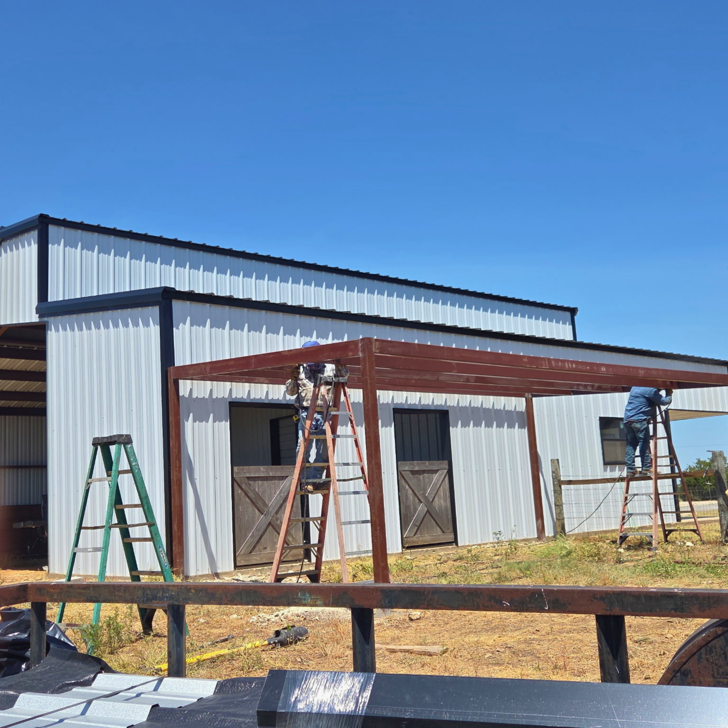 CMD Construction site with workers building a metal structure, ladders, wooden beams, and roofing materials under a clear blue sky.