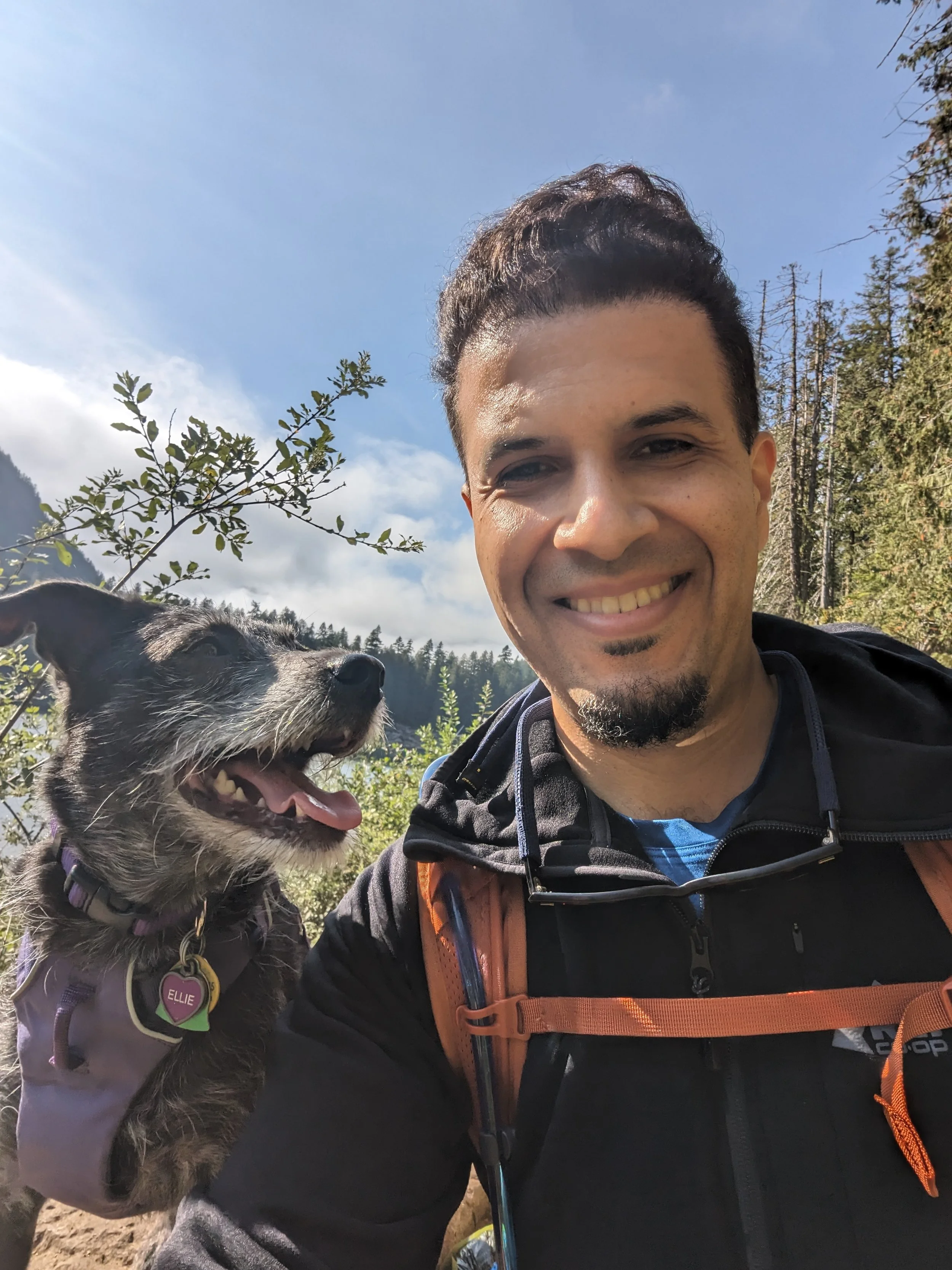 Dr. Correia smiling outdoors with a small black and grey dog wearing a purple harness and a pink heart-shaped tag. The background shows trees and a blue sky.